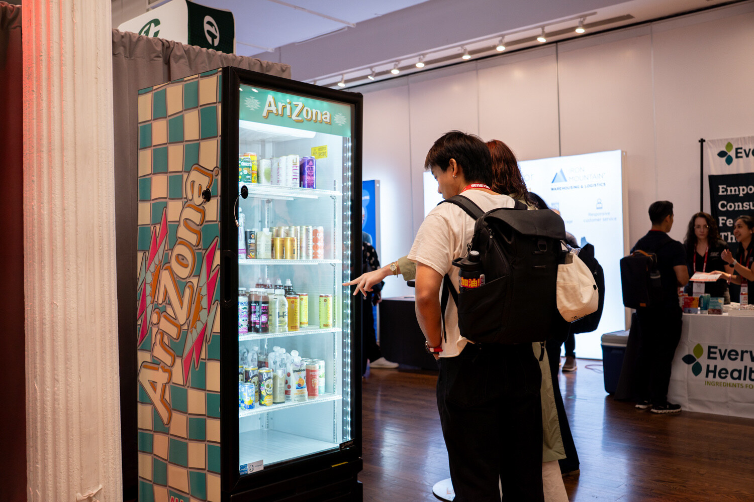 Two attendees with backpacks point into a lit fridge wrapped in AriZona’s checkered teal design.