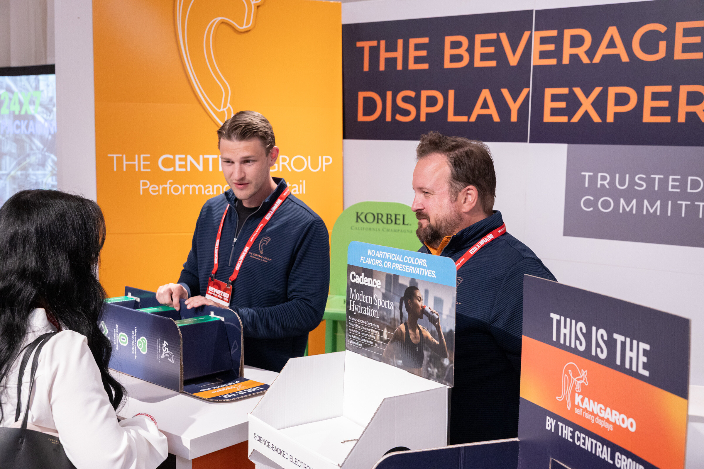 Young man in a navy jacket arranges a cardboard display while a colleague and an attendee look on, under an orange and navy The Central Group sign.