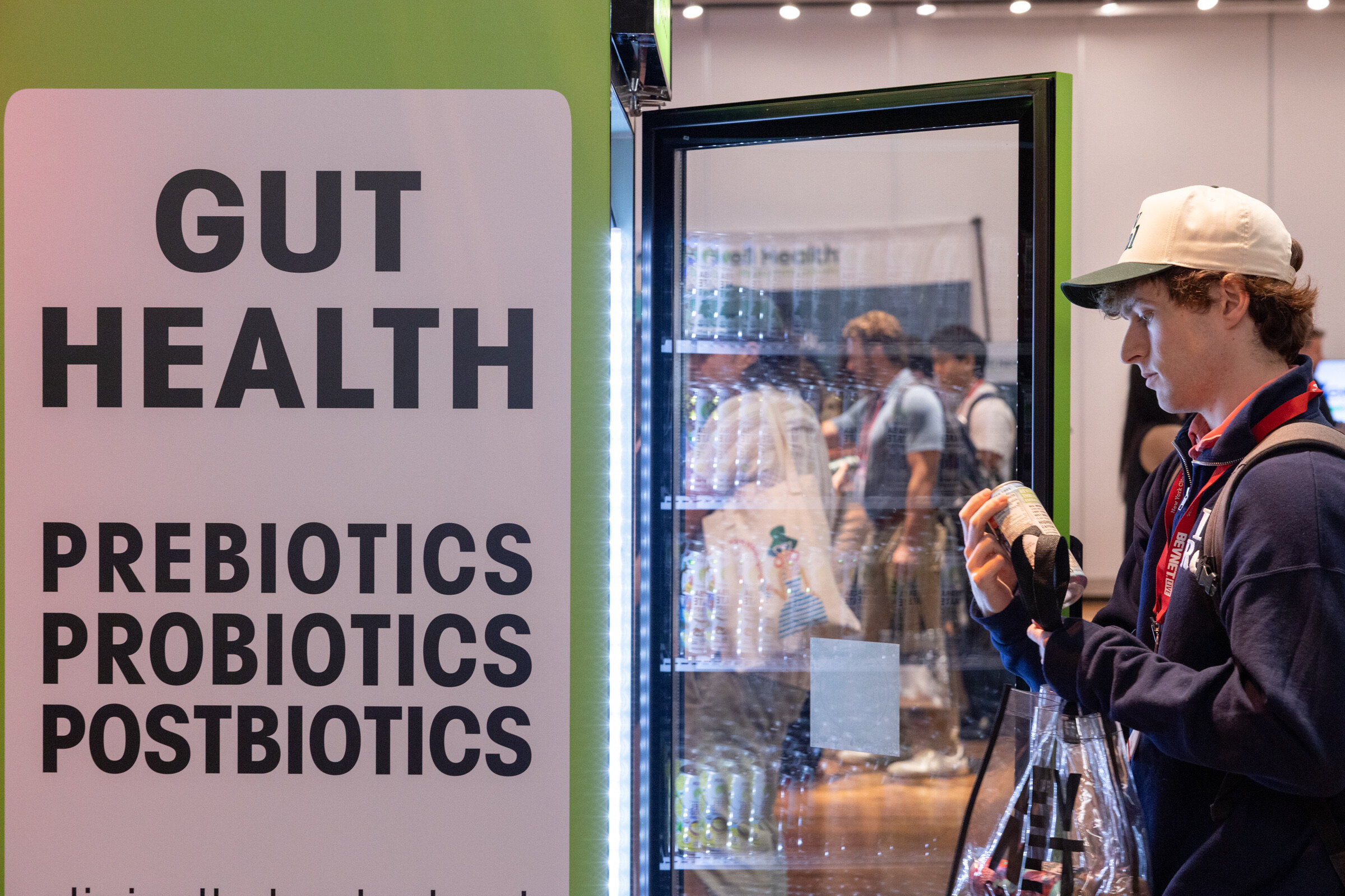 Young man in a cap holds a can beside a green fridge with a sign reading "Gut Health."