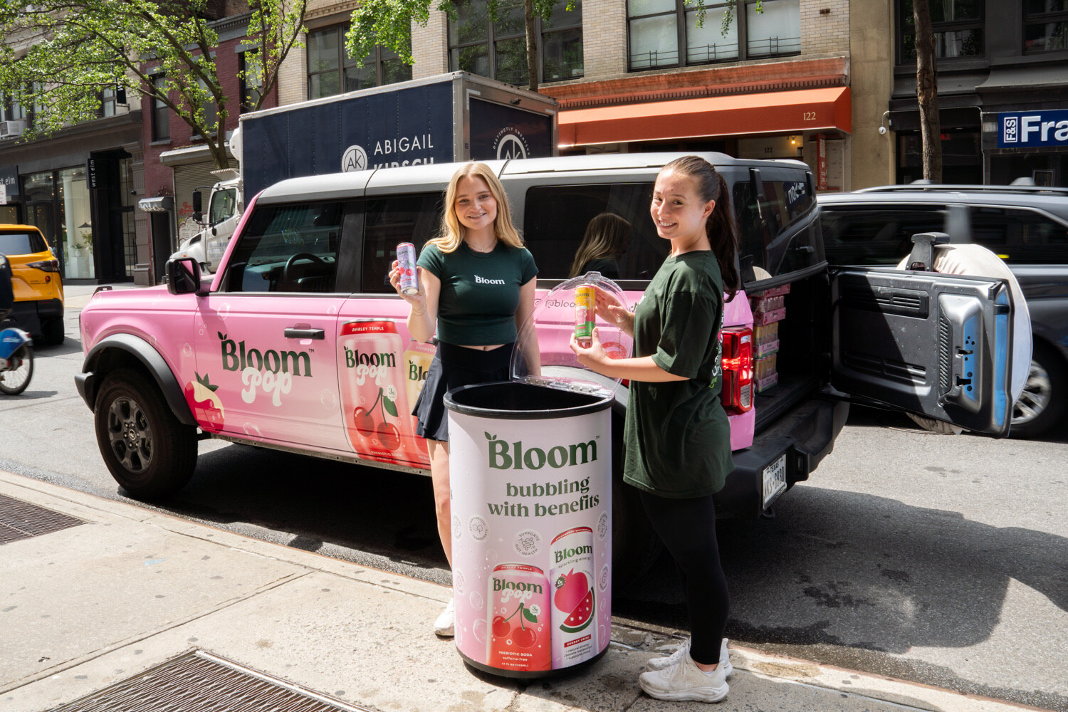 Two smiling women in green Bloom shirts hold cans beside a pink SUV wrapped in Bloom Pop graphics and a Bloom cooler.
