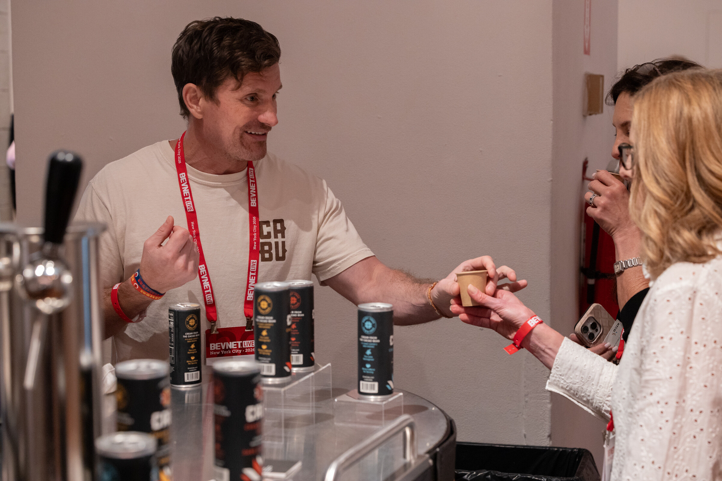 Smiling man in a CABU T-shirt hands a small cup to an attendee across a cart lined with CABU cans.