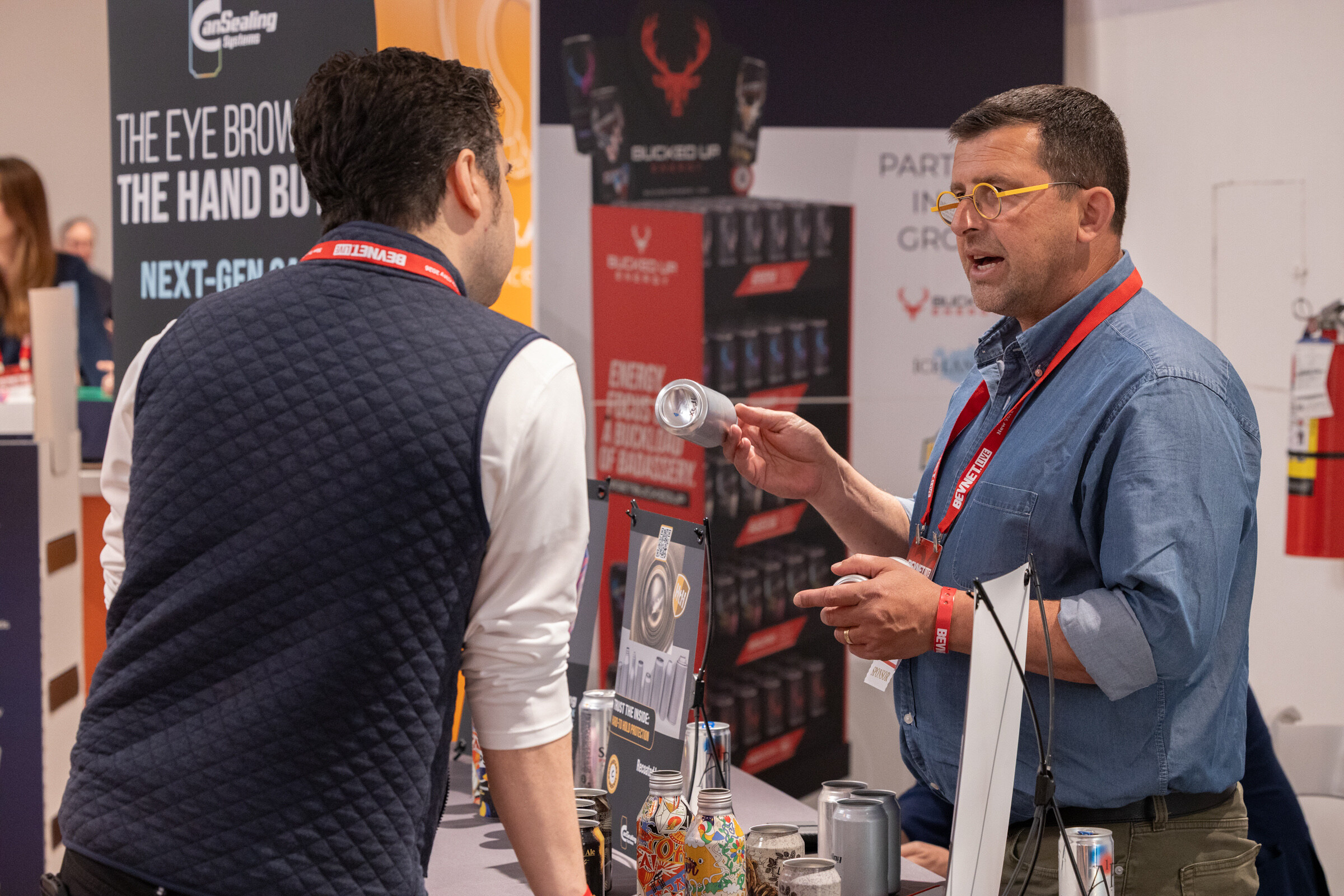 Man in yellow glasses and a denim shirt holds up an aluminum can while talking with an attendee in a quilted vest.