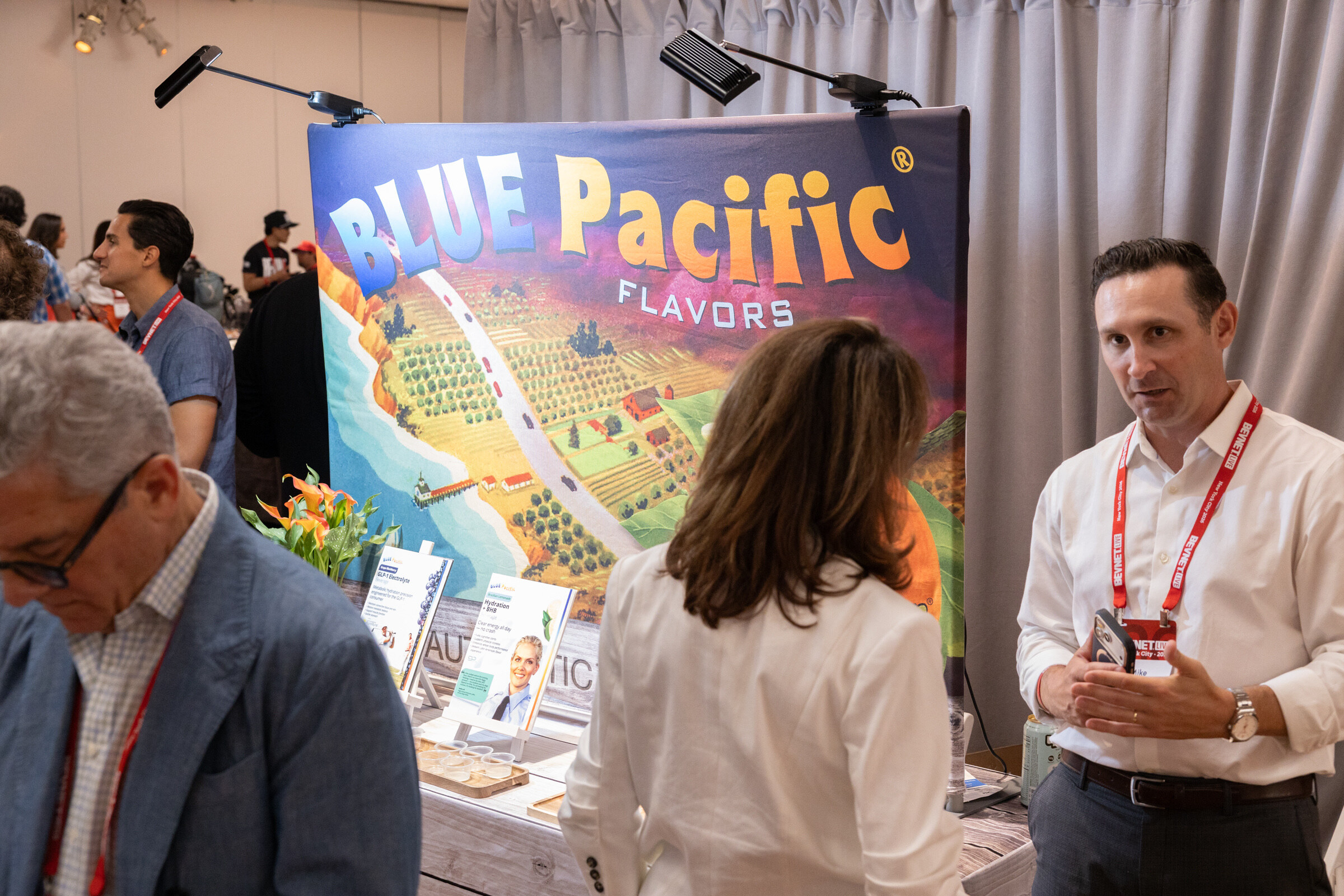 Man in a white shirt gestures while talking with two attendees beside the Blue Pacific Flavors backdrop.