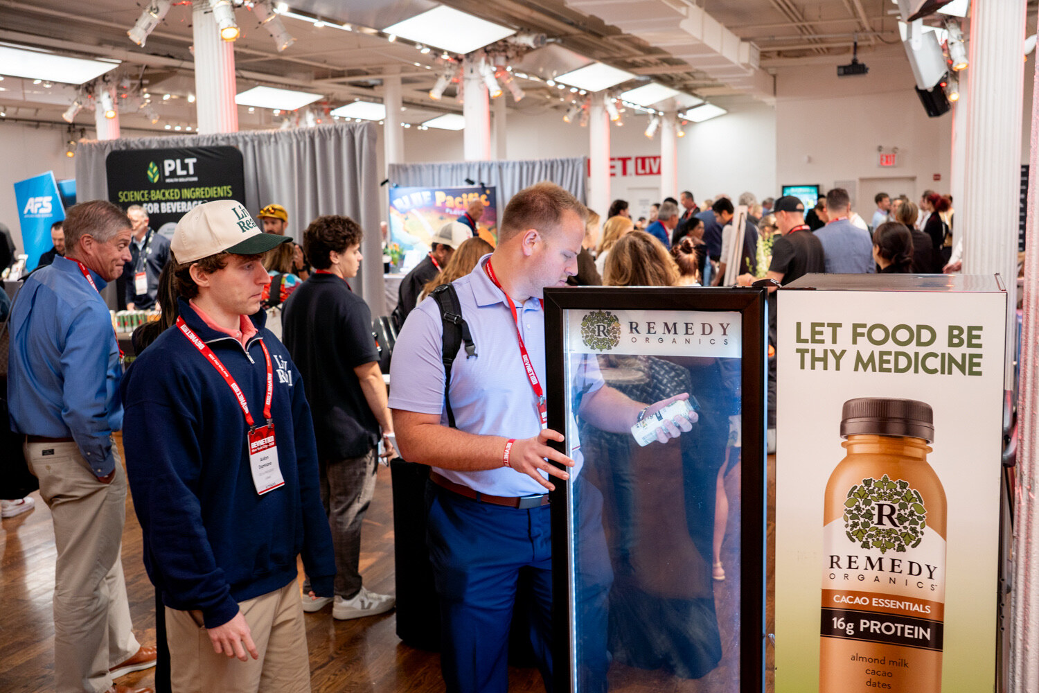 Man in a light blue shirt opens a Remedy Organics fridge beside a "Let food be thy medicine" sign in a crowded hall.