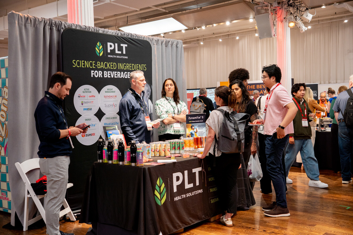 Crowd of attendees around a black PLT Health Solutions table and backdrop in a busy hall.
