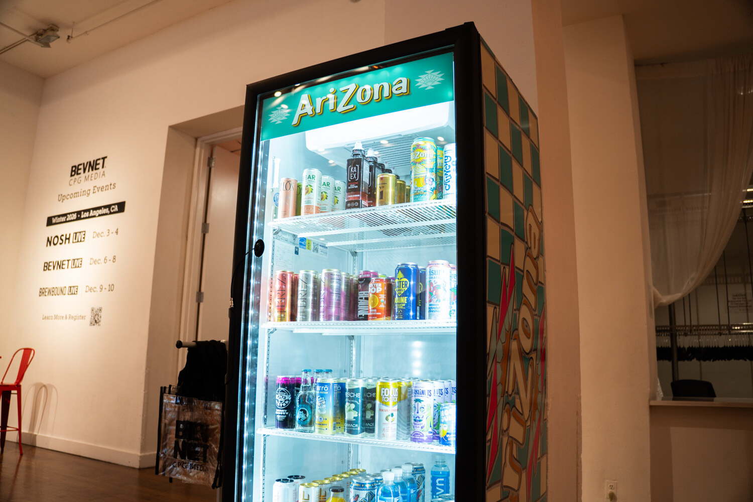 A lit glass-door fridge with an AriZona header and checkered teal side, full of cans and bottles, next to a wall of BevNET event graphics.