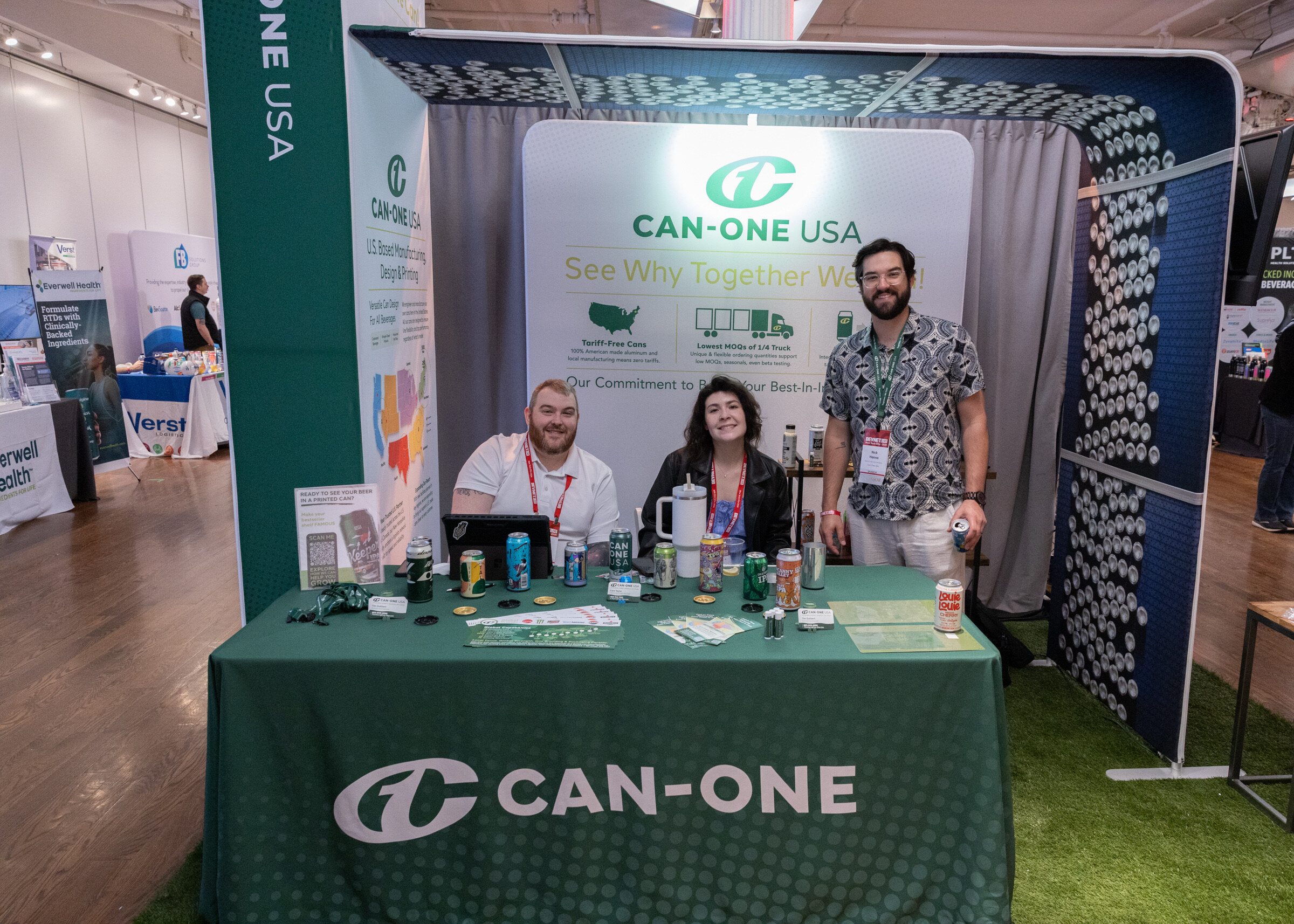 Three smiling people at a green Can-One table on artificial turf, with cans on display and a Can-One USA backdrop.