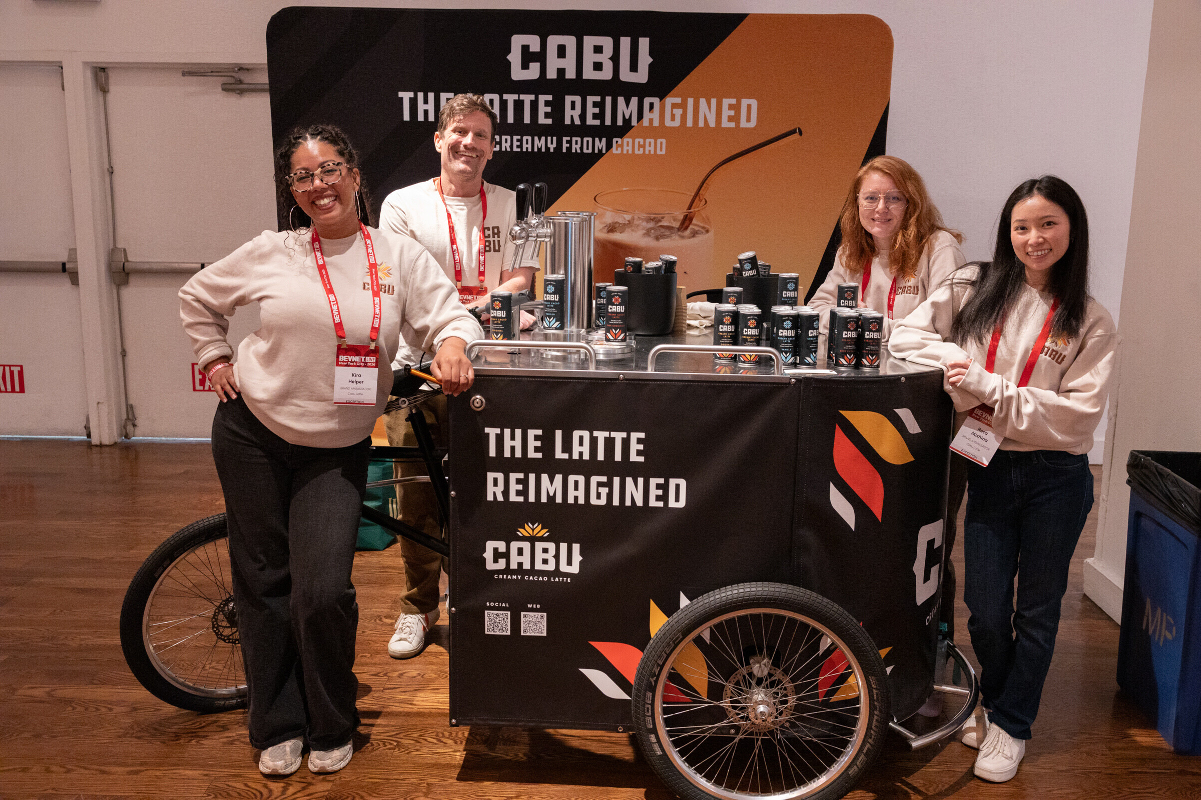 Four smiling people in CABU sweatshirts stand around a black tricycle cart labeled "The Latte Reimagined" in front of a CABU backdrop.