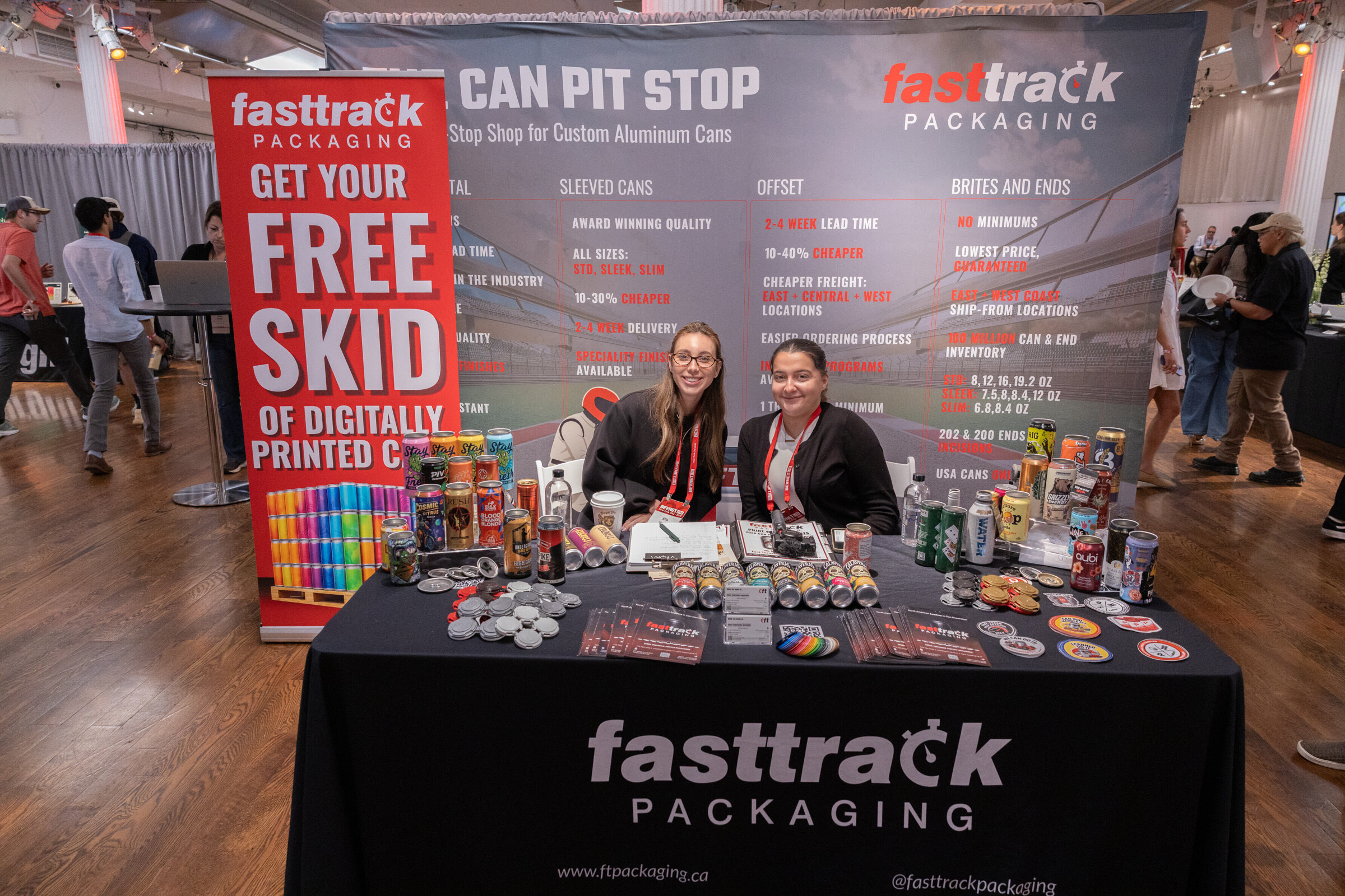 Two smiling women sit behind a black Fasttrack Packaging table covered with printed cans, next to a "Get your free skid" banner.