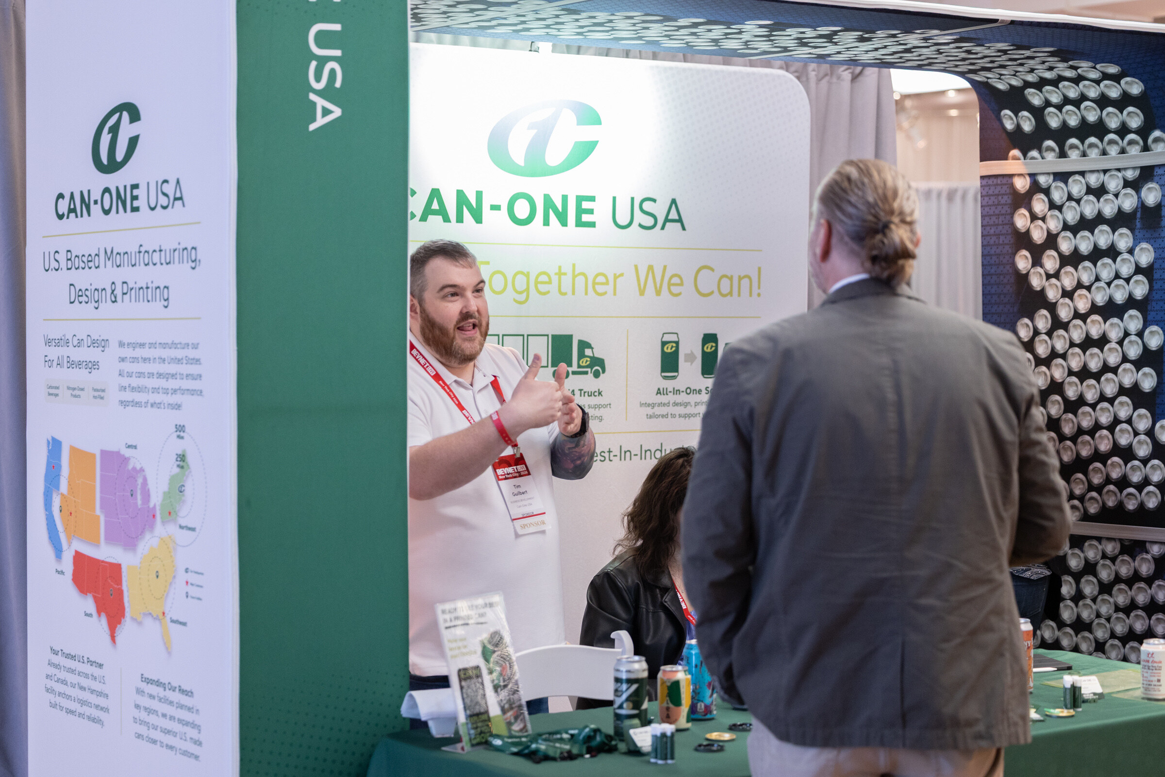 Man in a white shirt gives a thumbs-up while talking with an attendee in a gray jacket at the Can-One USA booth.
