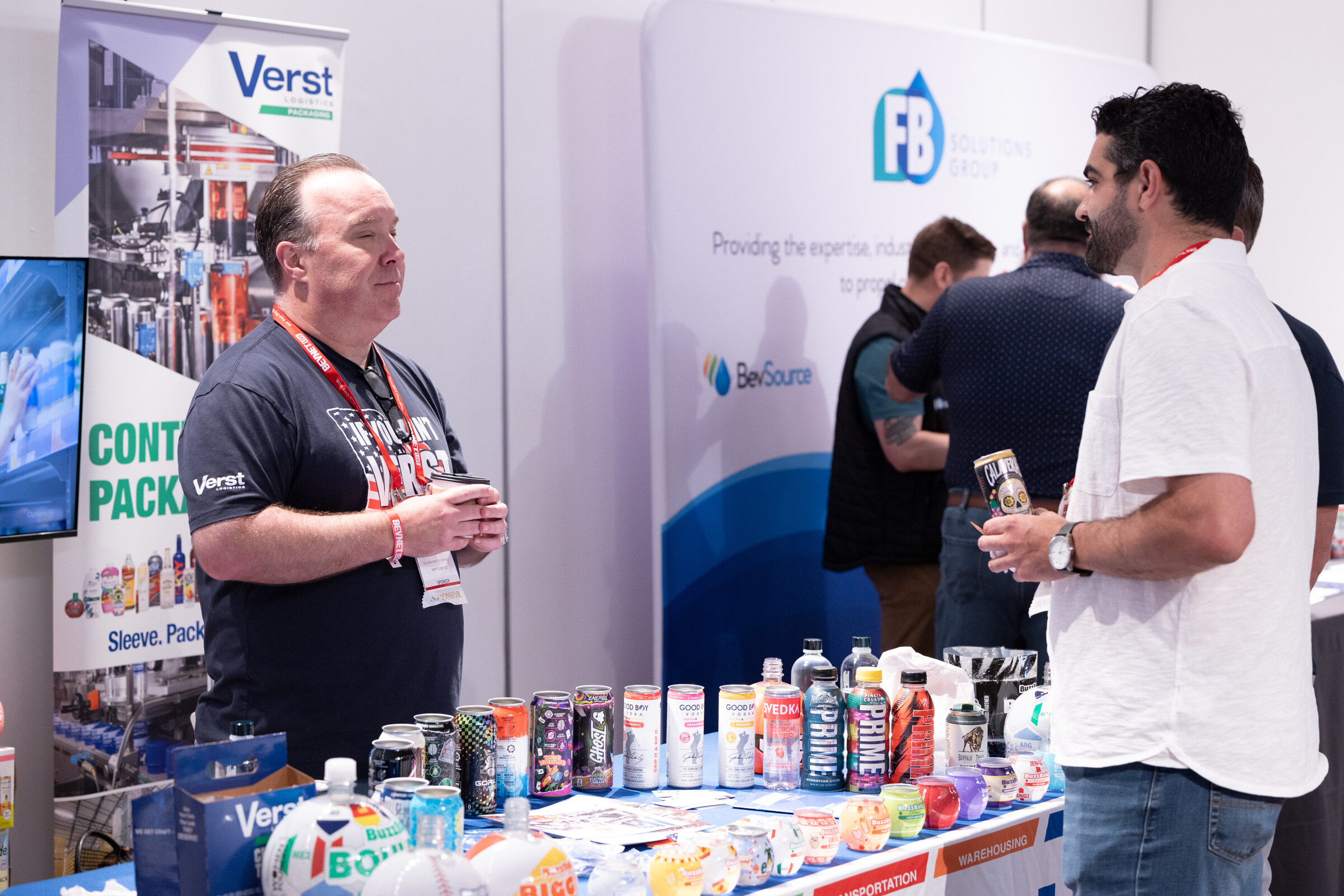 Man in a Verst T-shirt holding a coffee stands behind a table of colorful drinks while attendees talk nearby.