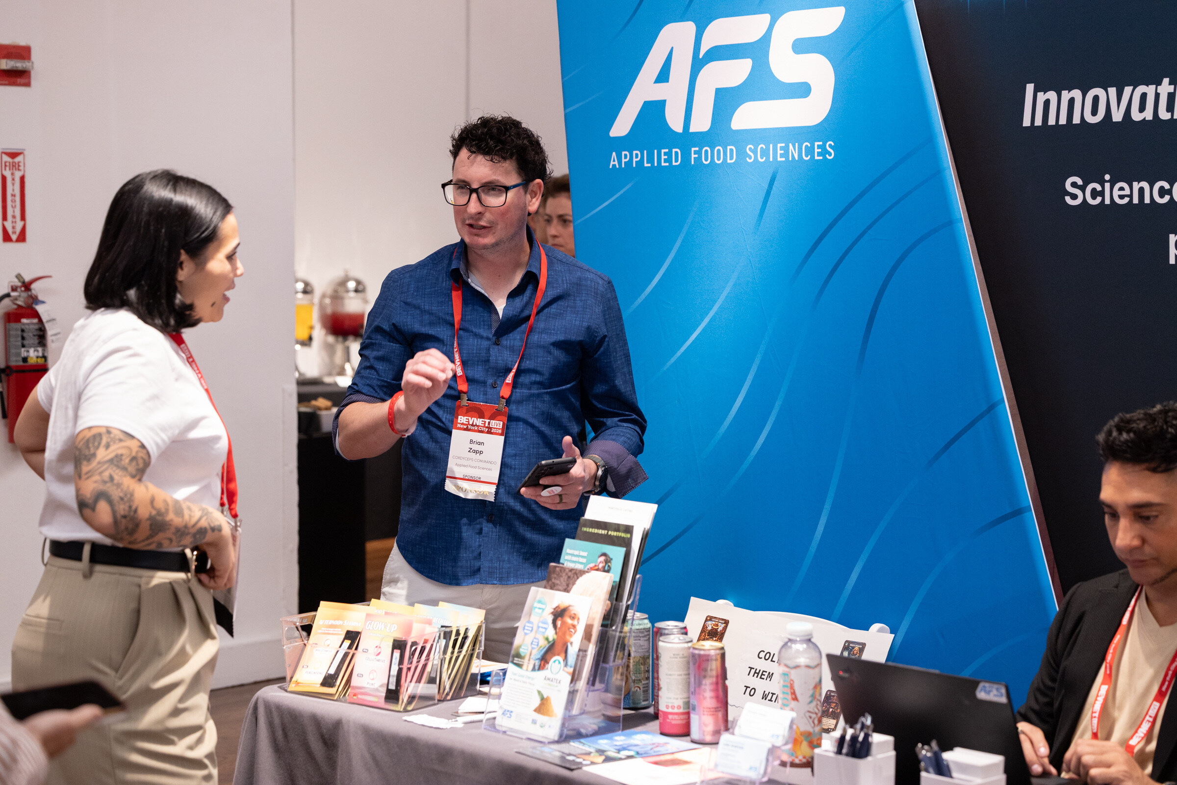 Man in glasses and a blue shirt talks with a woman in a white T-shirt beside a blue AFS banner and a table of brochures.