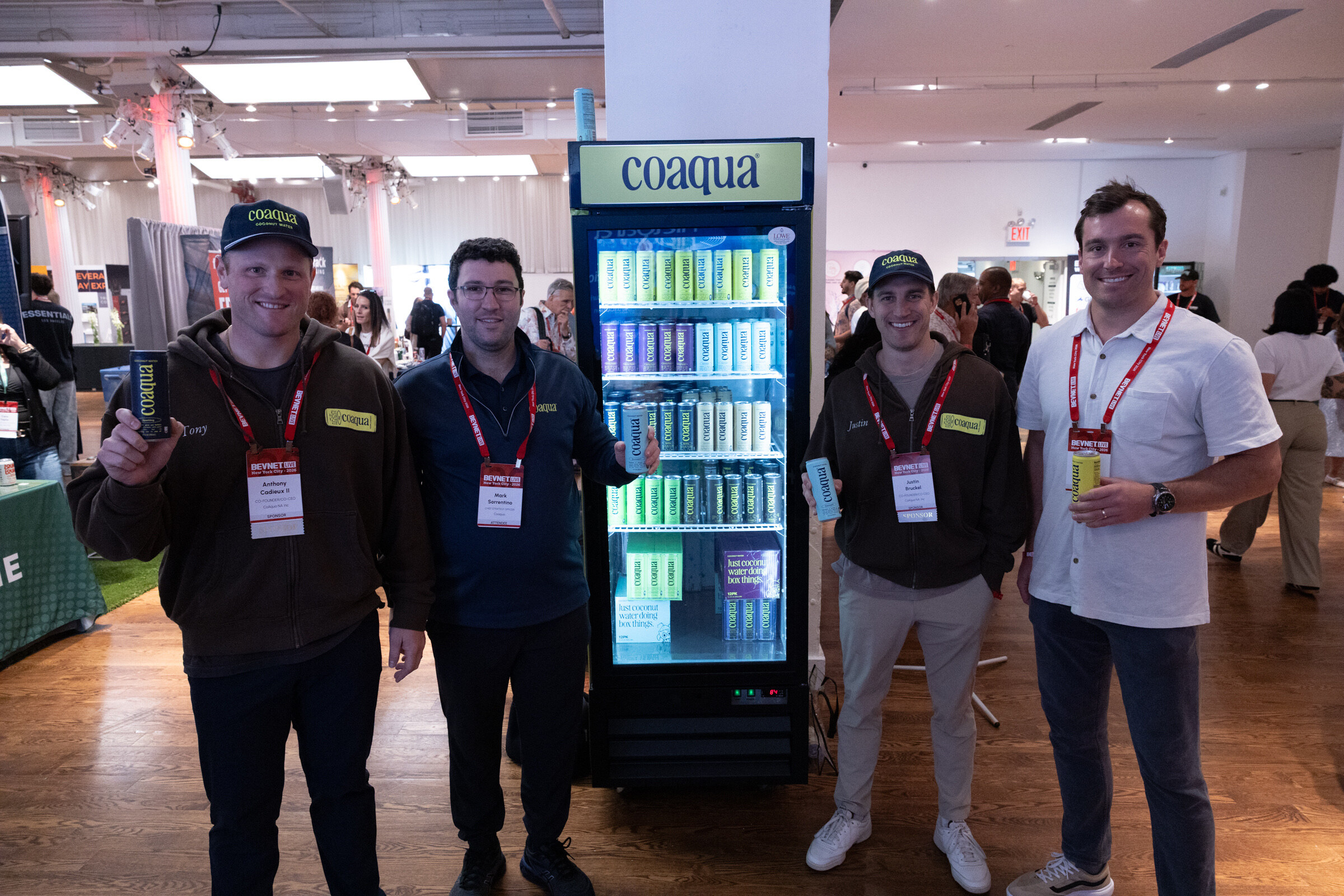Four smiling men, two in coaqua caps holding cans, stand on either side of a lit coaqua fridge.