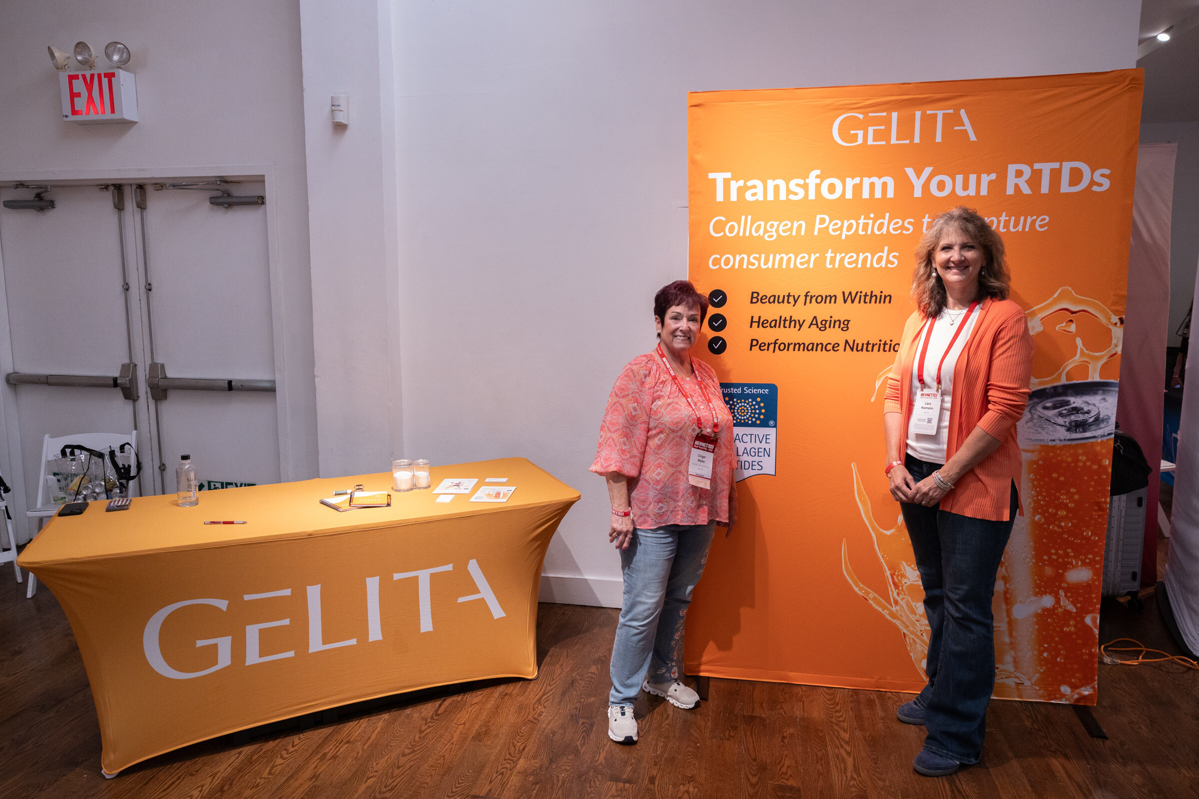 Two smiling women stand beside an orange GELITA banner reading "Transform Your RTDs" and an orange GELITA table.