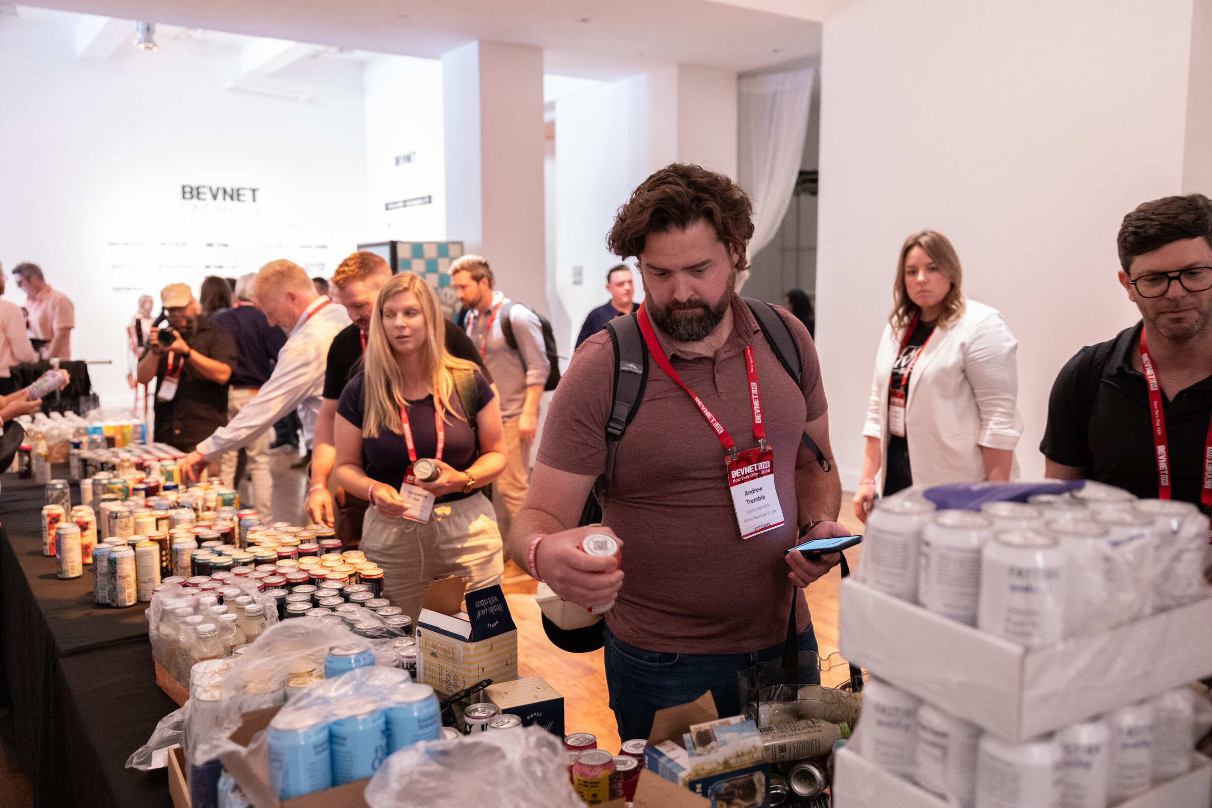 Attendees browse a long table crowded with cans and bottles, one man holding a bottle and his phone.