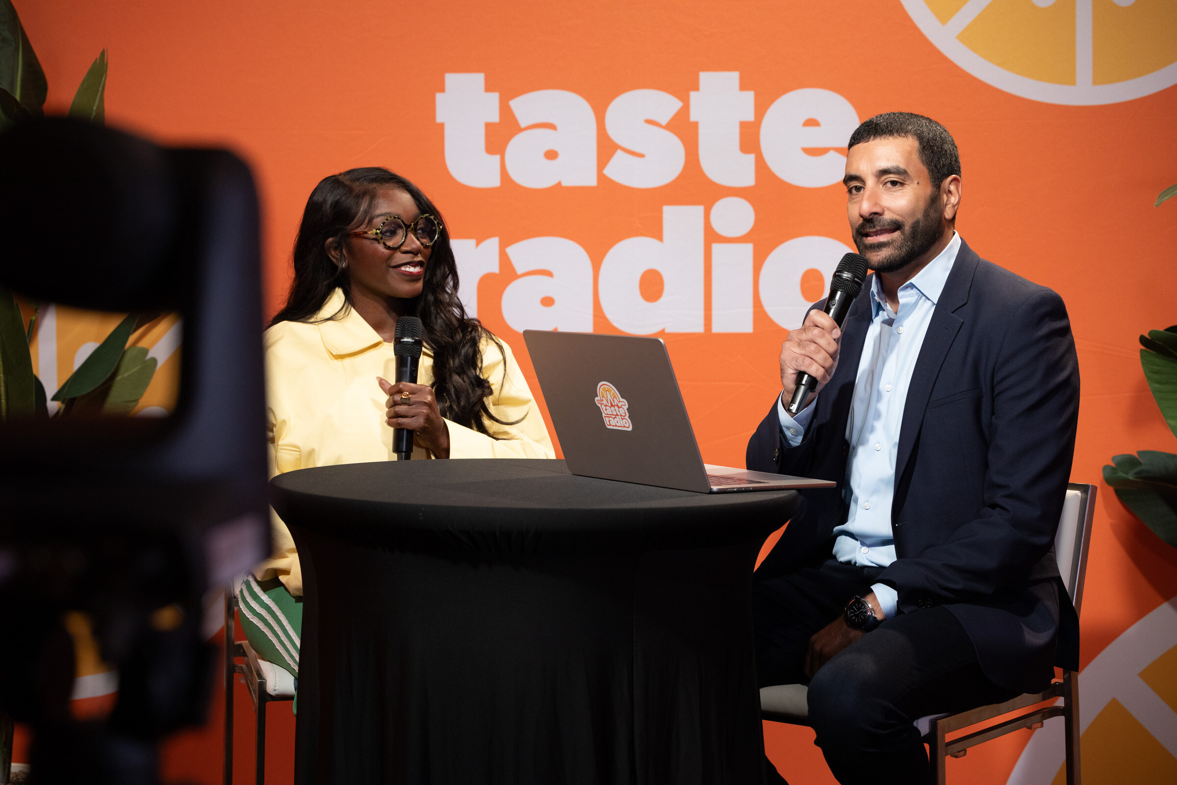 Woman in a yellow jacket and glasses and a man in a navy suit with microphones at a high-top table in front of the Taste Radio backdrop.