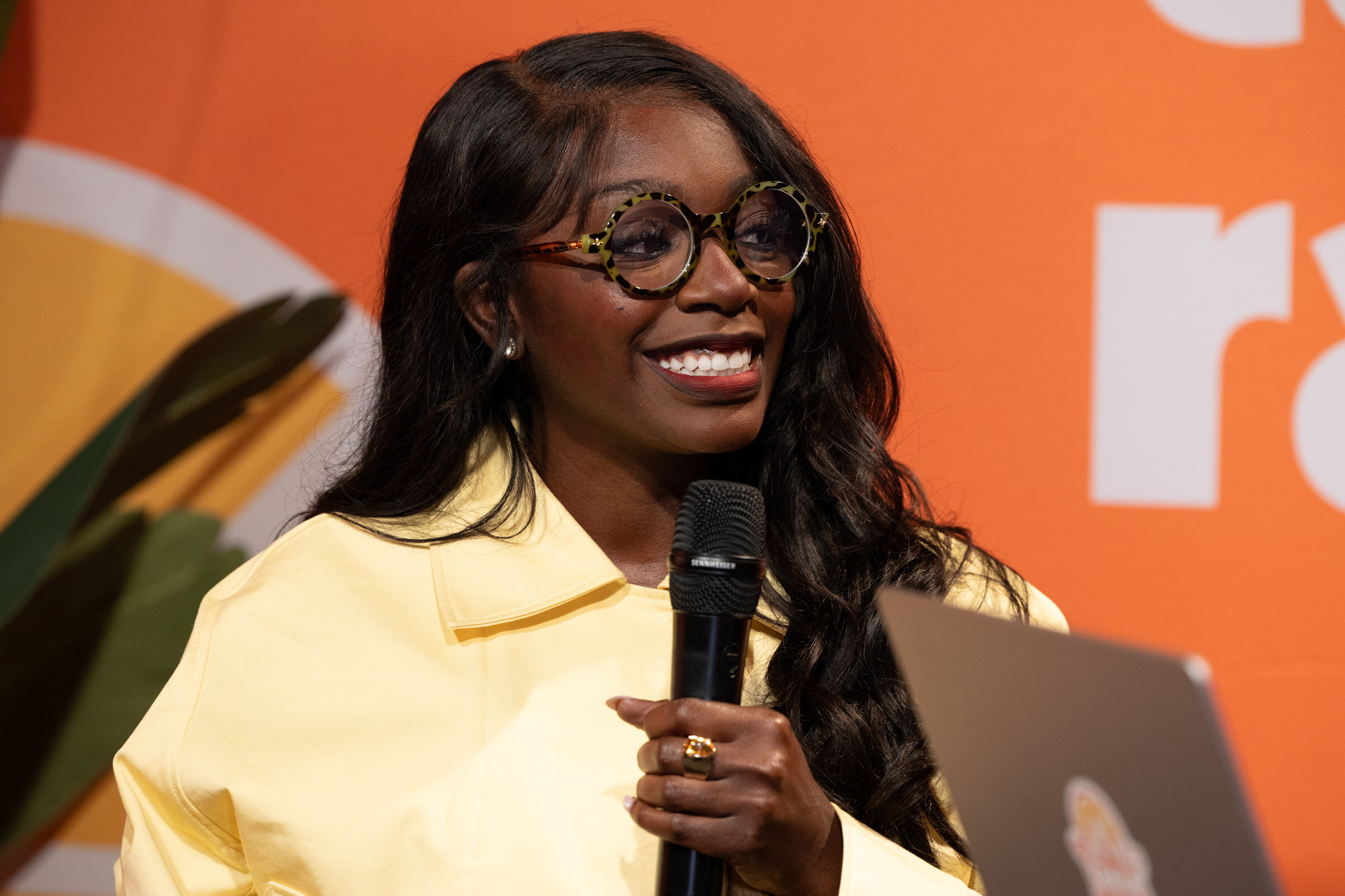 Smiling woman in tortoiseshell glasses and a yellow jacket holding a microphone in front of an orange backdrop.