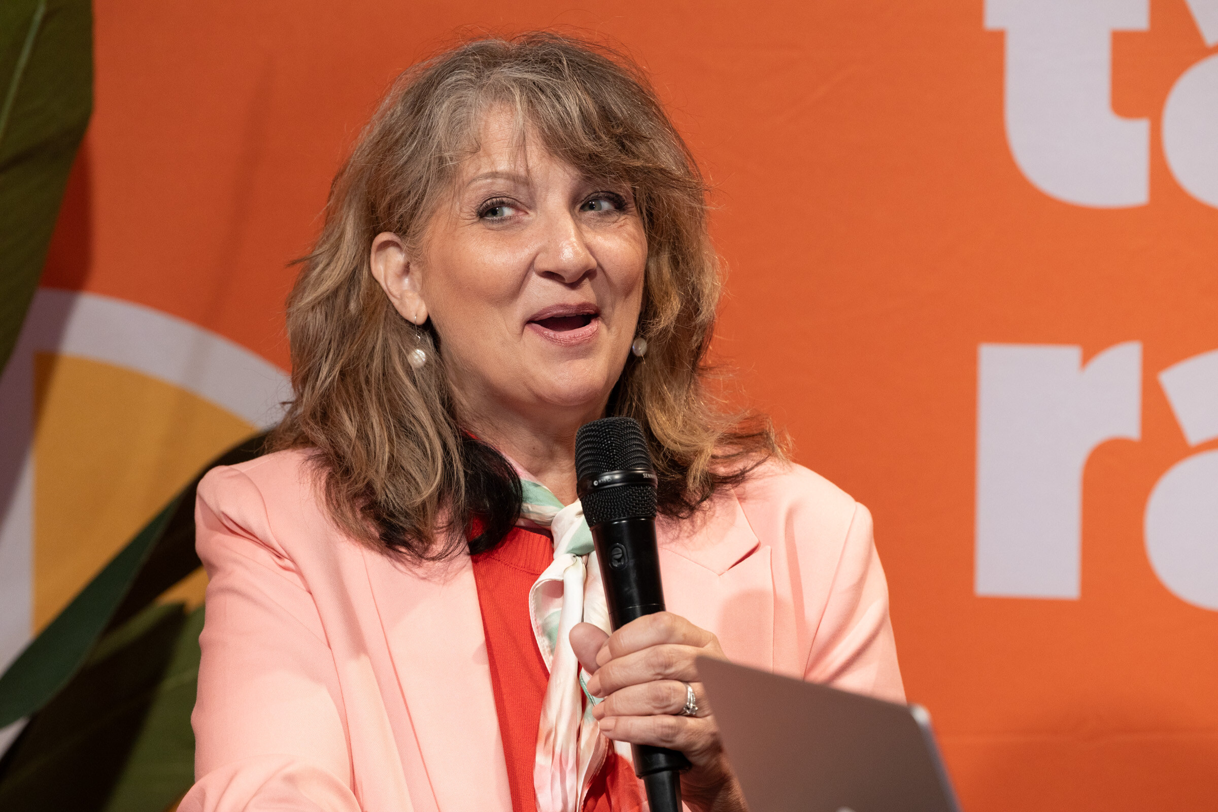 Woman with wavy hair in a pink blazer and patterned scarf speaking into a microphone.