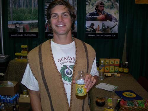 A smiling man in a tan woven vest and a Guayaki Yerba Mate T-shirt holds a bottle of the drink at a booth with conservation photos and products on tables.