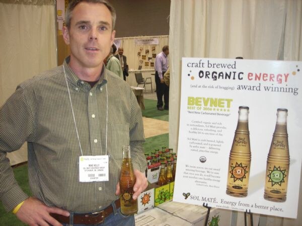 Mike Kelly of Sol Maté Beverage Group stands in a green plaid shirt holding a bottle of Sol Maté. Beside him is a poster reading craft brewed organic energy with a BevNET Best of 2006 mention.