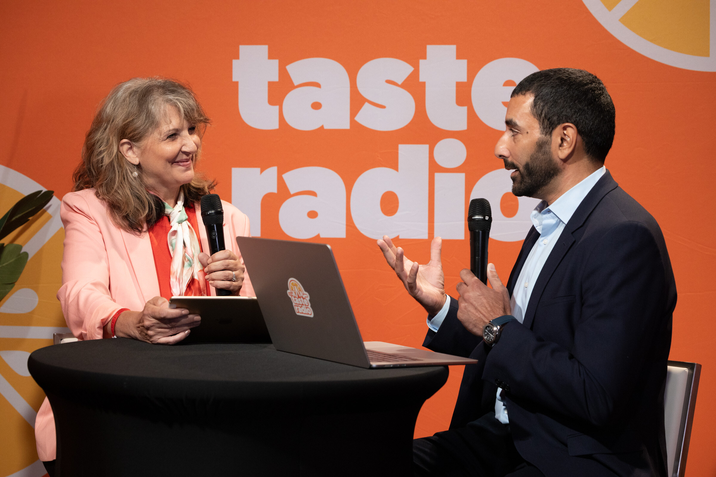 Woman in a pink blazer and a man in a navy suit talking at a high-top table with a laptop, in front of the Taste Radio logo.