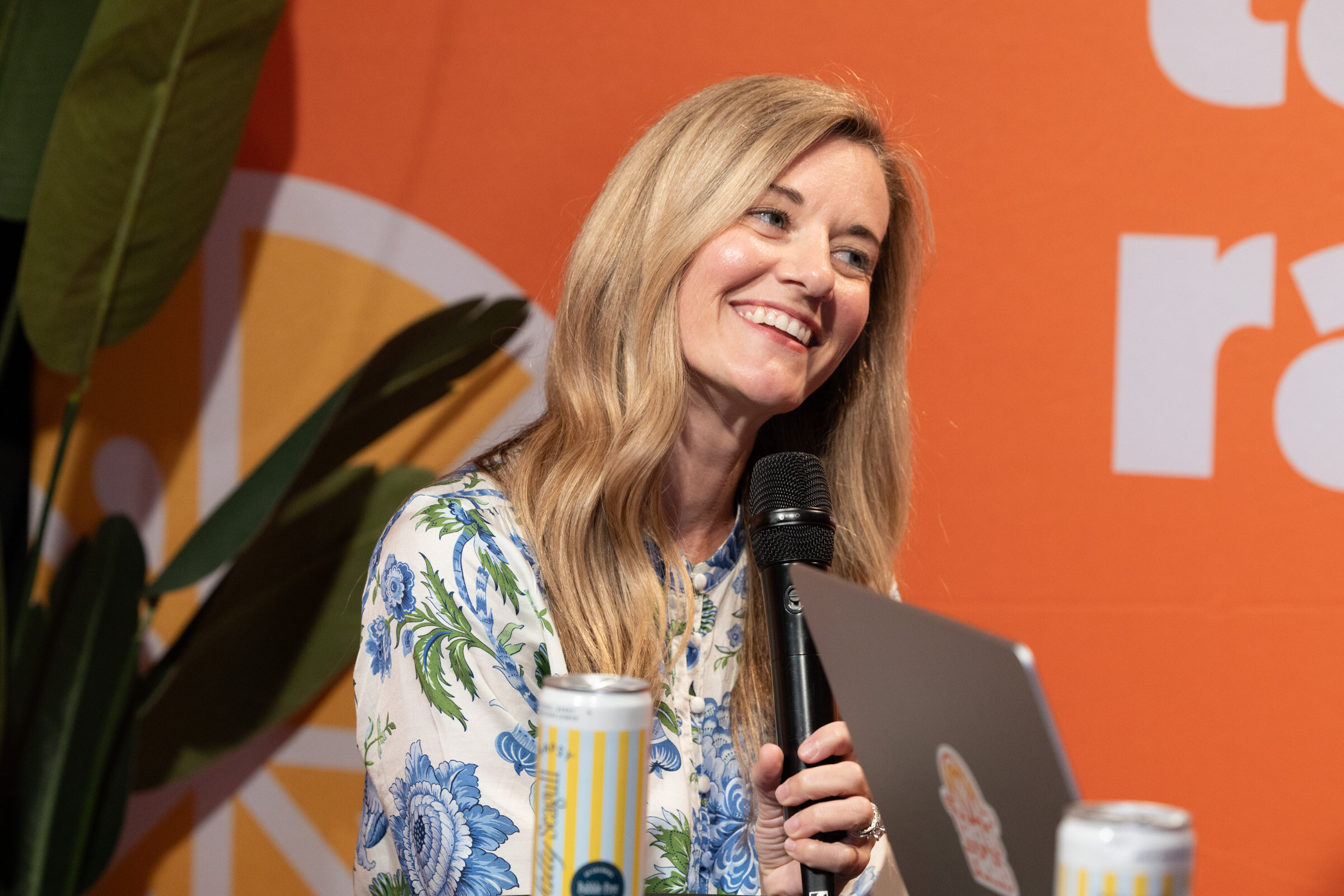 Smiling woman with long blond hair in a blue floral blouse holding a microphone, a can on the table in front of her.