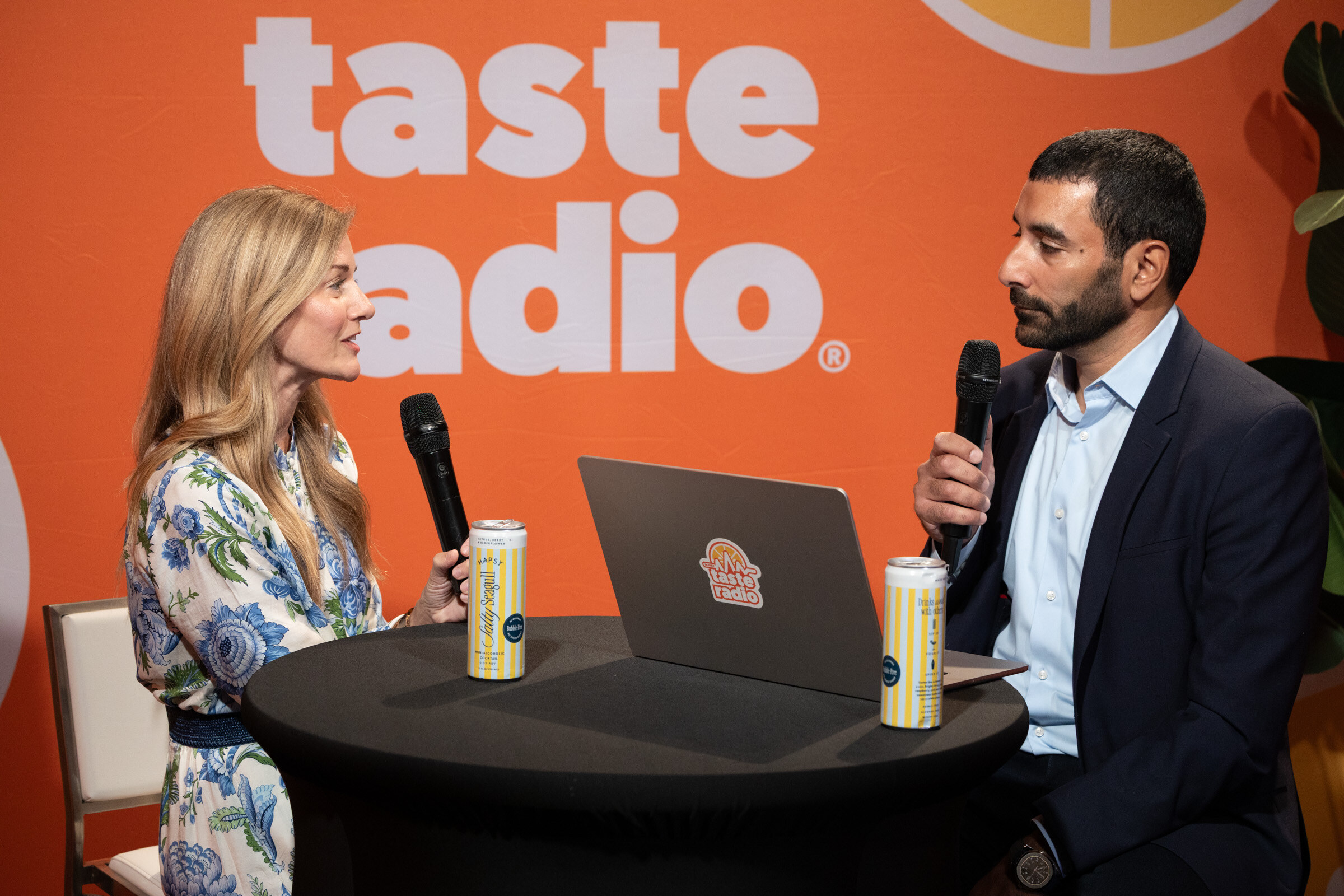Woman in a floral blouse and a man in a navy suit with microphones at a high-top table with two cans and a laptop.