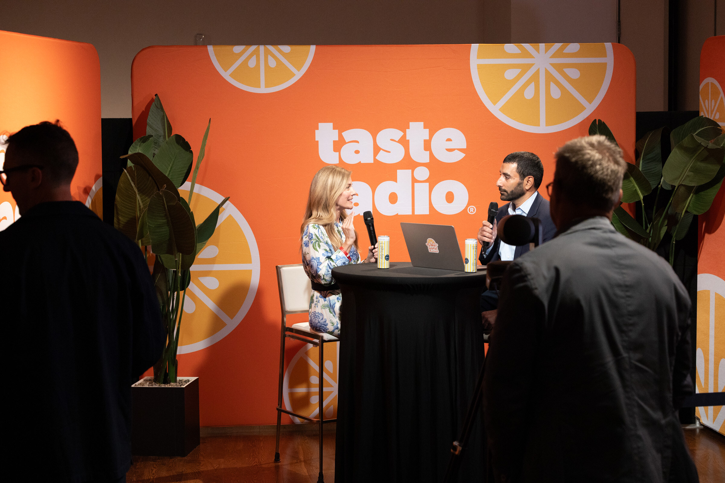 Two people seen from behind framing a Taste Radio interview at a high-top table in an orange studio set.