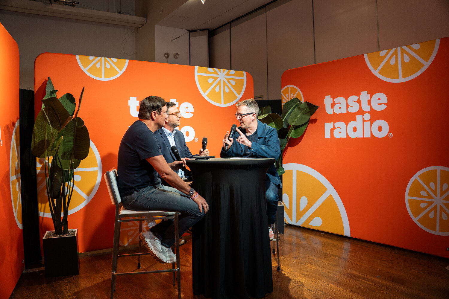 Three men talking at a high-top table in a studio set with orange Taste Radio walls, citrus graphics and potted plants.