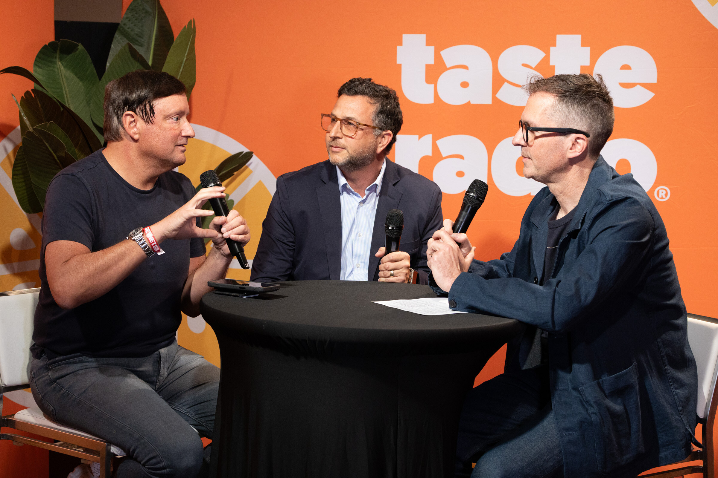 Three men with microphones talking around a high-top table in front of the Taste Radio backdrop.