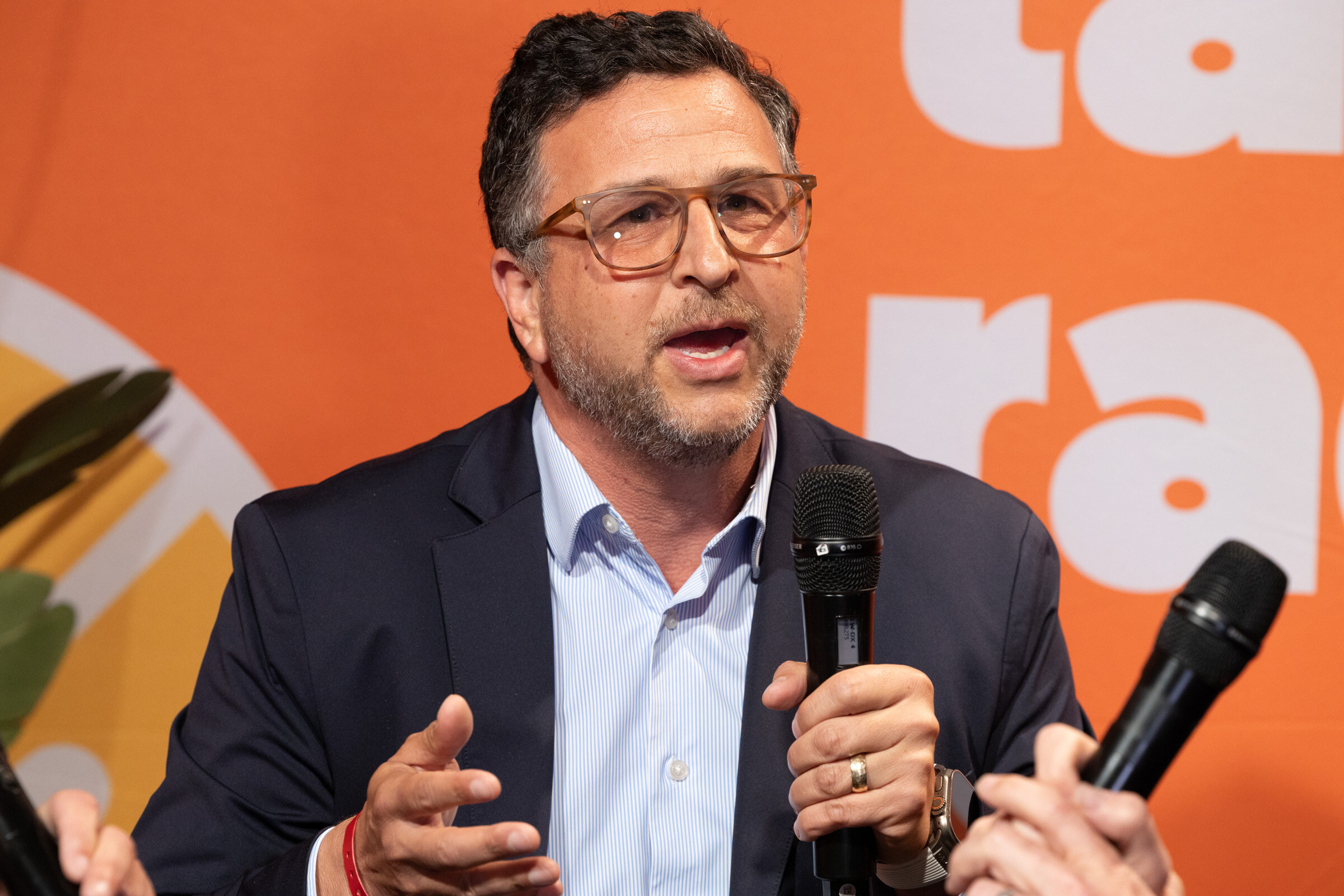 Man in glasses and a navy blazer speaking into a microphone and gesturing, in front of an orange backdrop.