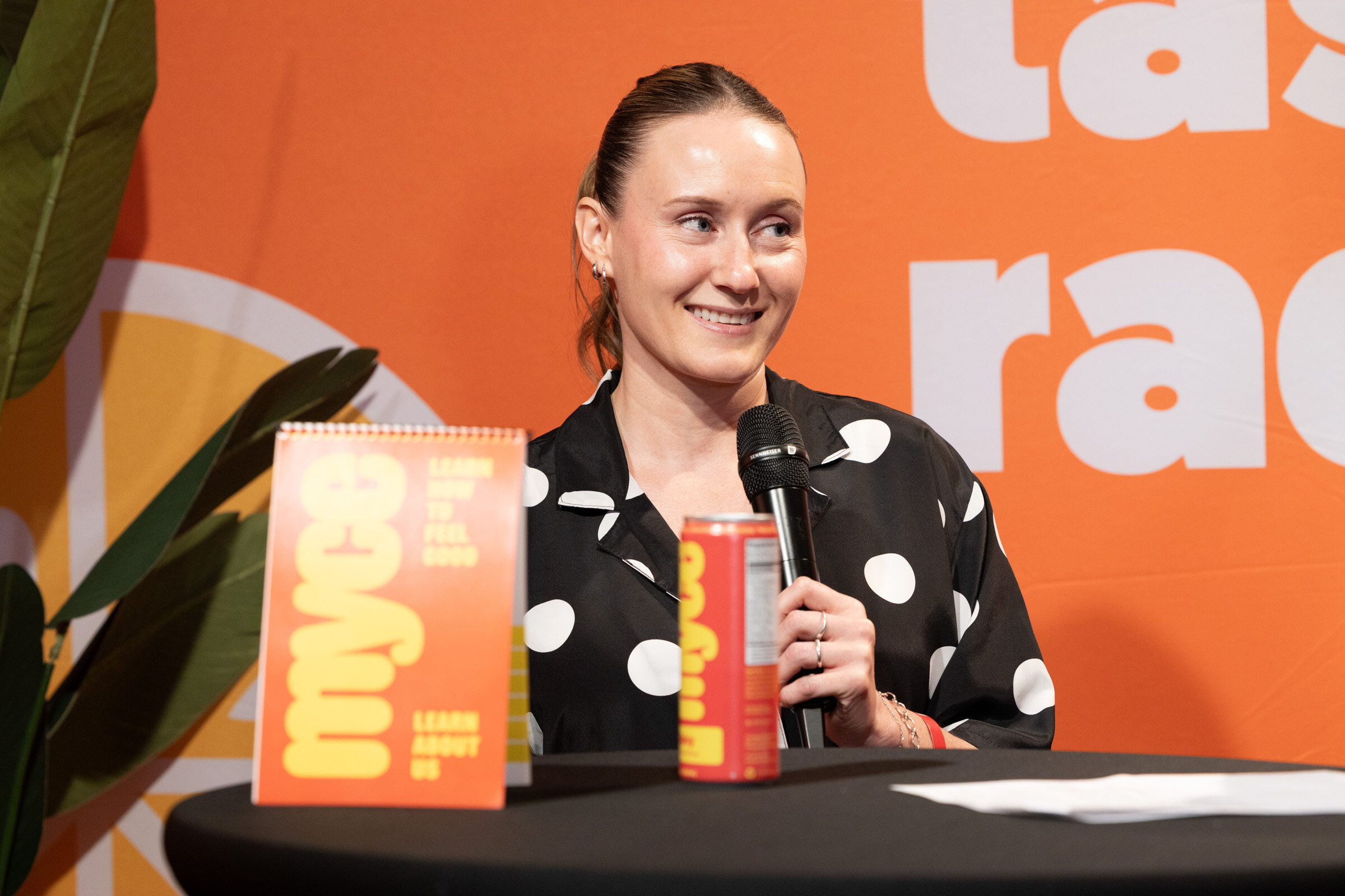 Smiling woman in a polka-dot shirt holding a microphone behind an orange Myce sign and a red Myce can.
