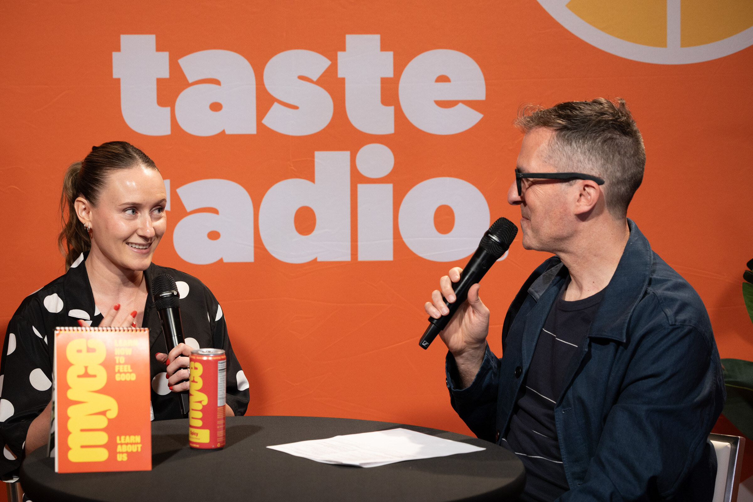 Woman in a polka-dot shirt smiling at a man in glasses speaking into a microphone, with a Myce sign and can on the table.