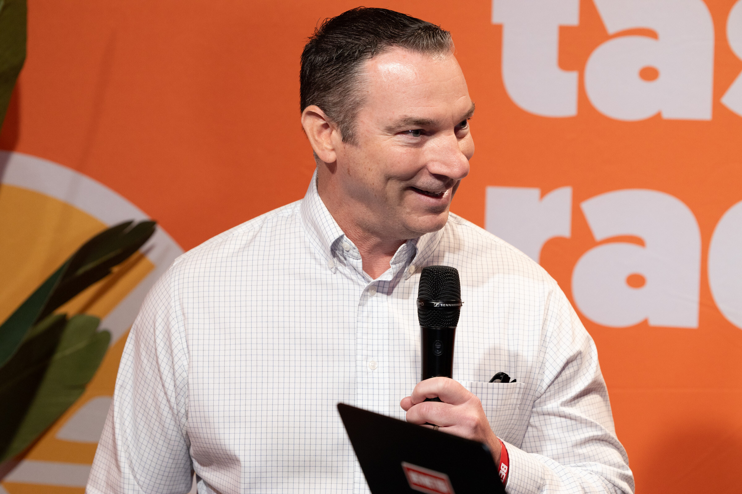 Smiling man in a white checked shirt holding a microphone and a tablet in front of an orange backdrop.