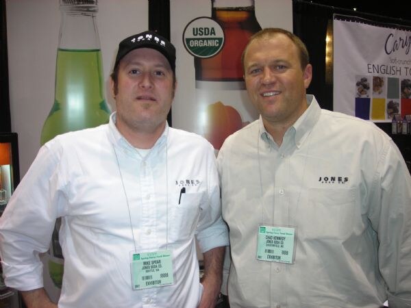 Mike Spear, left, in a black cap, and Chad Kennedy, right, both of Jones Soda Co., stand side by side in white shirts with Jones logos and exhibitor badges, in front of Jones Soda bottle graphics.