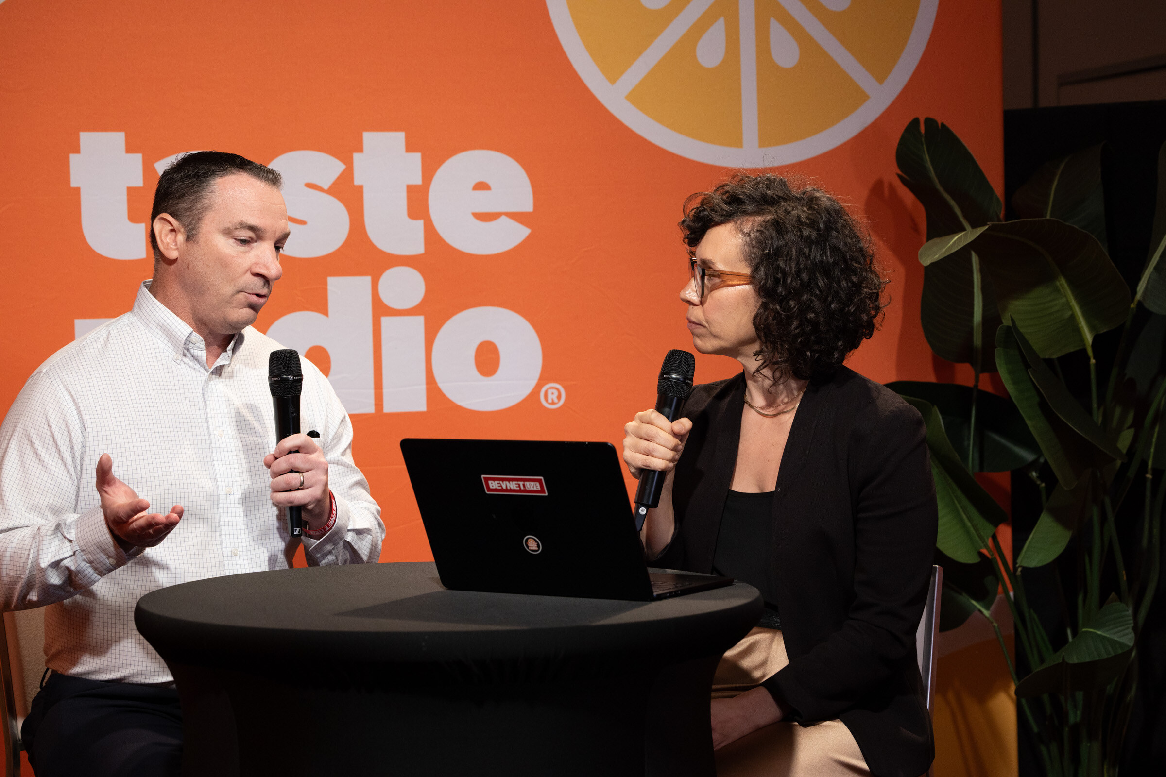 Man in a white shirt gesturing while talking with a woman with curly hair and glasses at a high-top table.