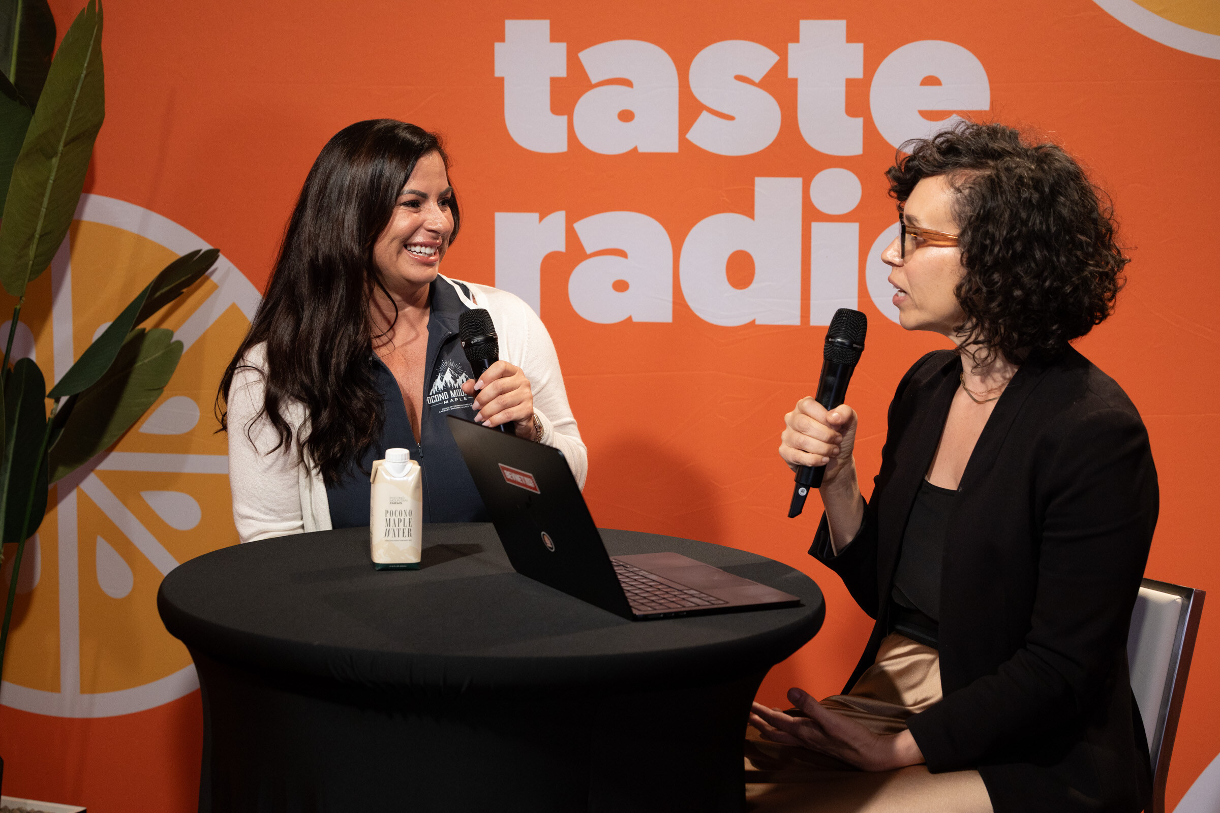 Woman with long dark hair in a Pocono Mountain Maple jacket smiling at a woman in glasses across a high-top table.