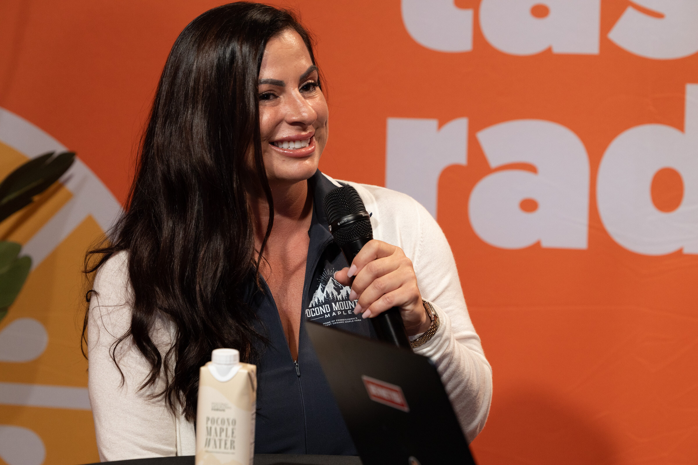 Smiling woman in a Pocono Mountain Maple jacket holding a microphone beside a carton of Pocono Maple Water.