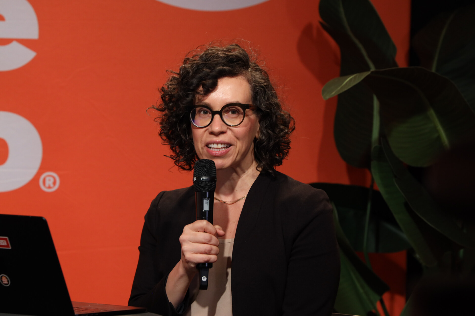 Woman with curly hair and round glasses speaks into a microphone in front of the orange Taste Radio backdrop and a potted plant.