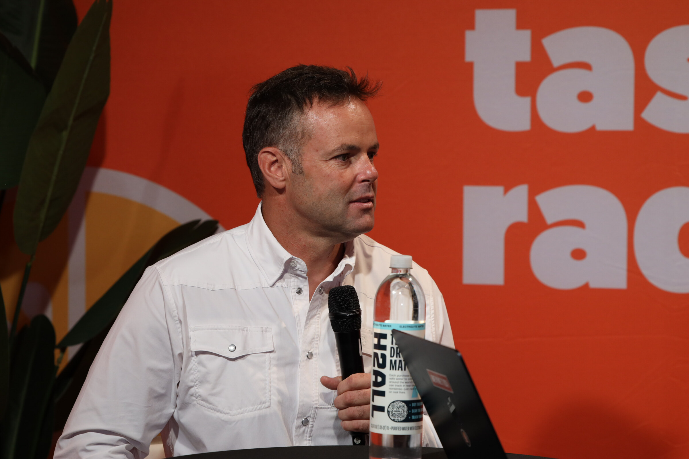 Man in a white western shirt holding a microphone next to a bottle of H2ALL water in front of an orange backdrop.