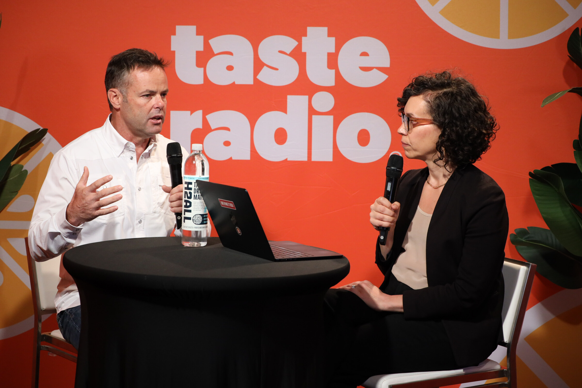 Man in a white shirt gesturing while a woman in glasses and a black blazer listens, with a bottle of H2ALL on the table.