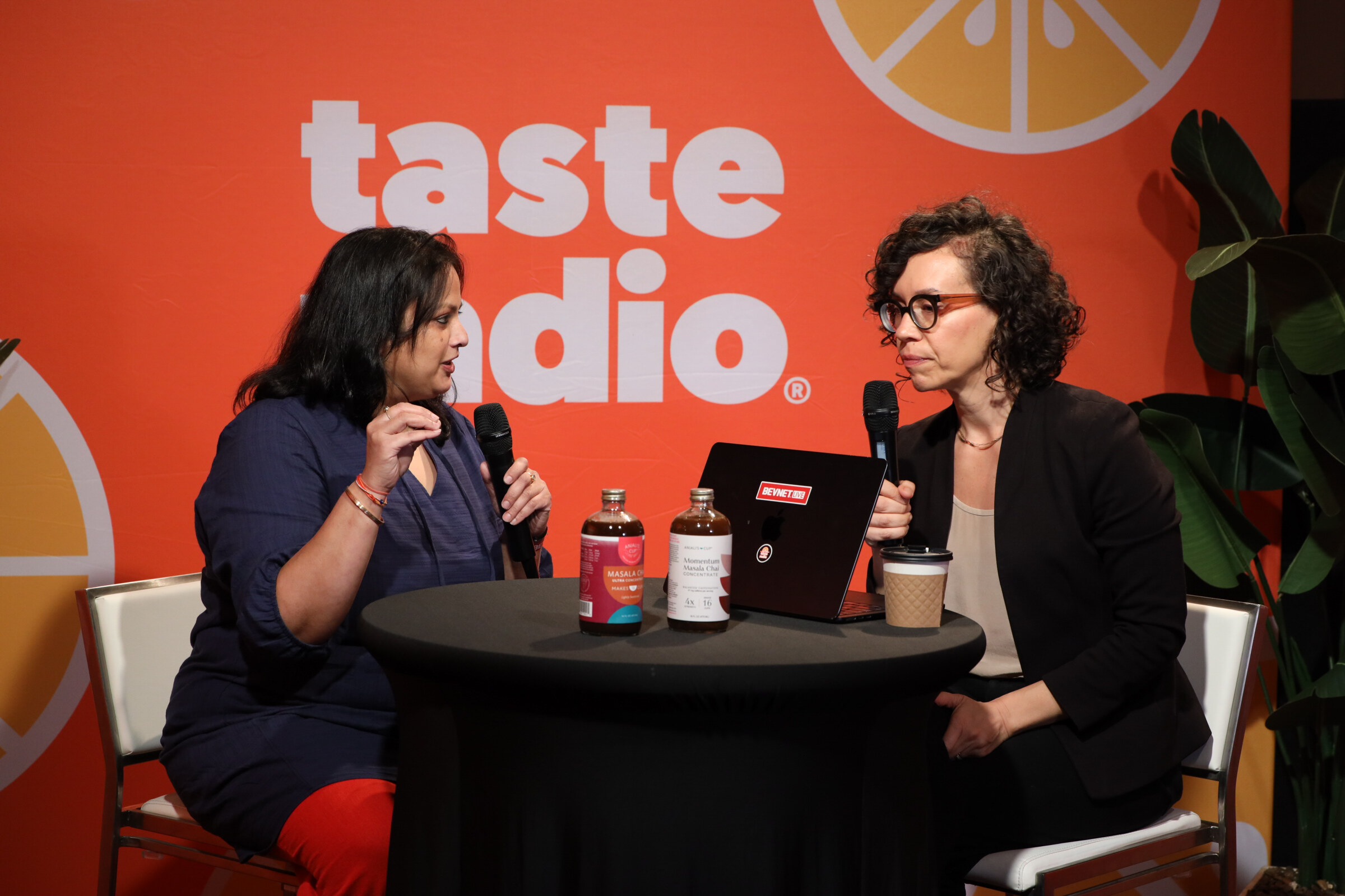 Woman in a navy dress talking with a woman in glasses at a high-top table with two bottles of masala chai concentrate and a coffee cup.