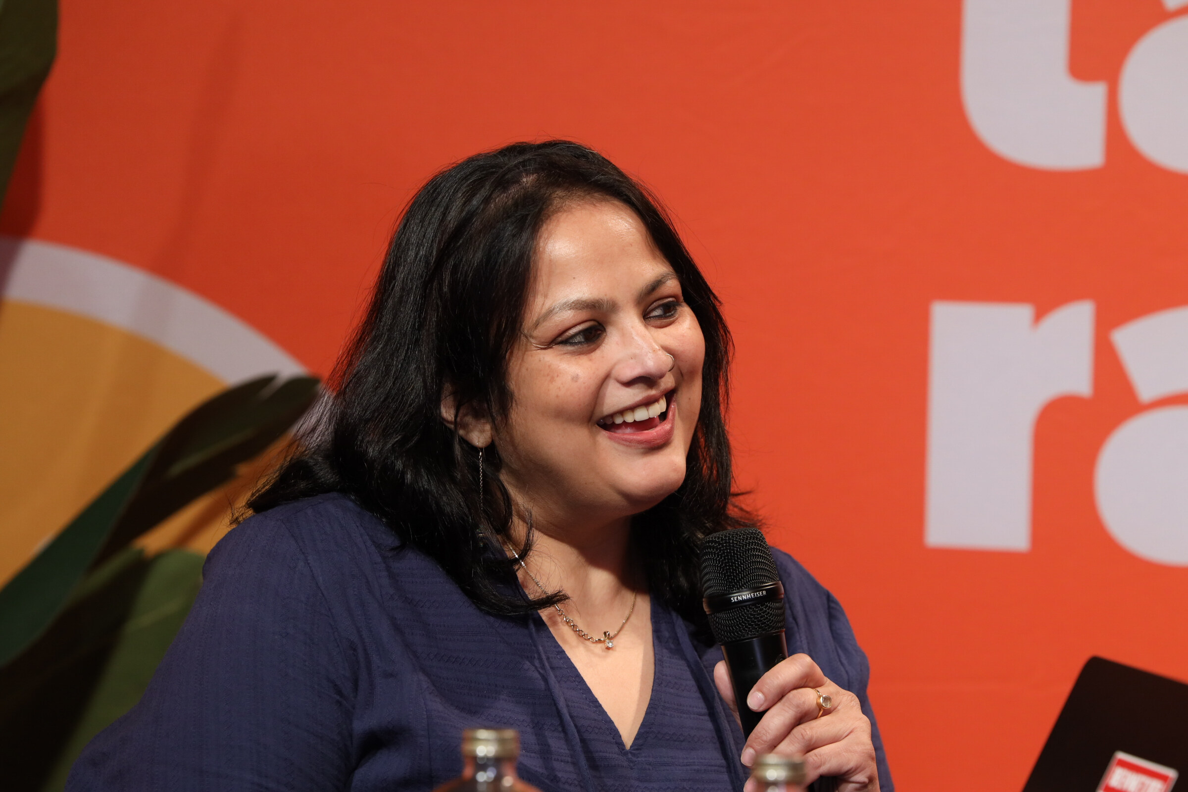 Laughing woman with dark hair in a navy top holding a microphone in front of an orange backdrop.