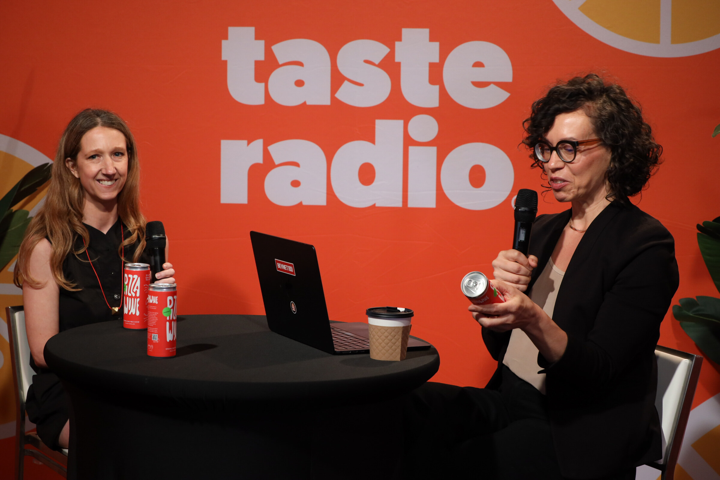 Woman in glasses and a black blazer holding a red can and microphone across a high-top table from a smiling guest with two red cans.