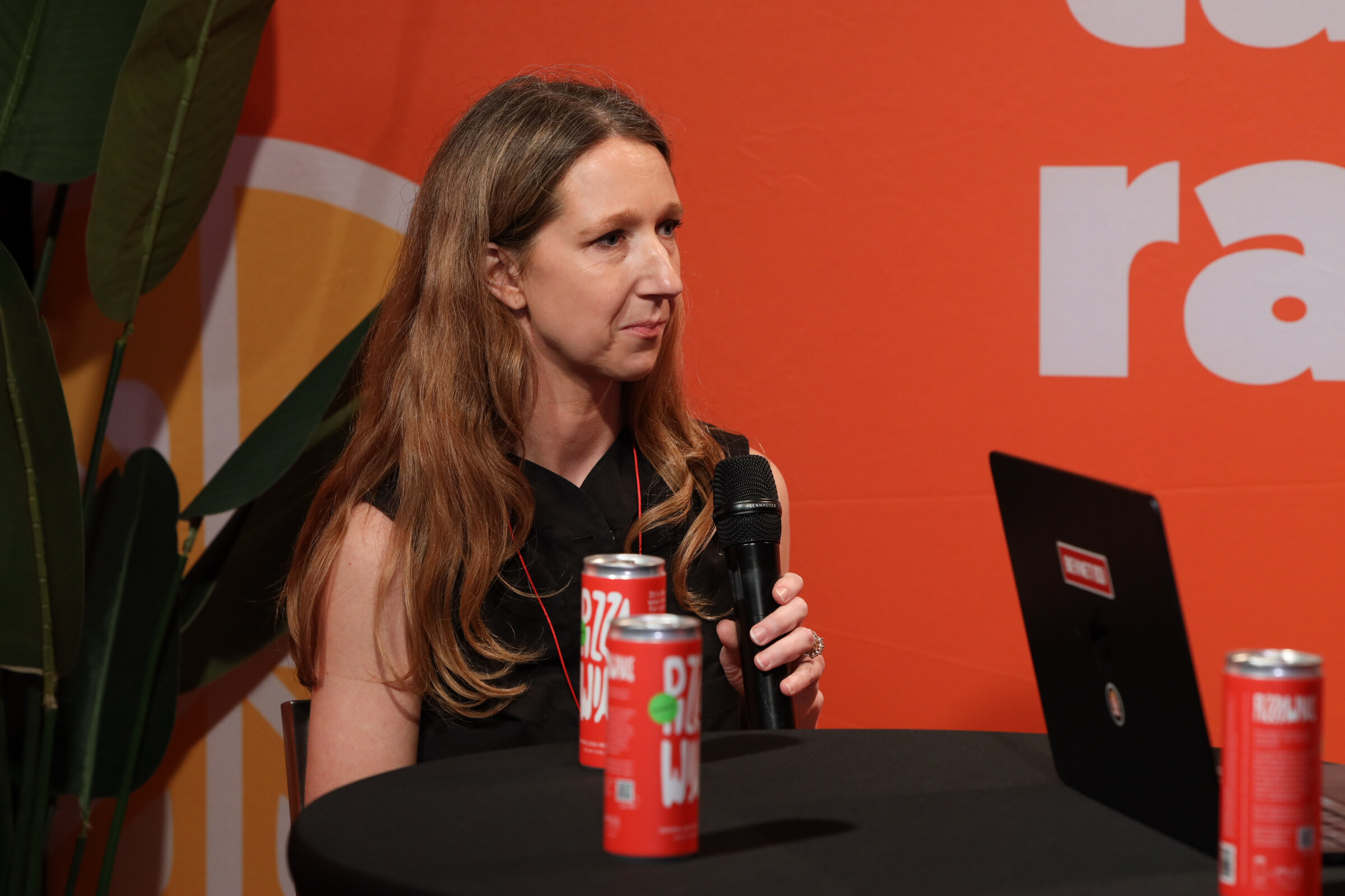 Woman with long hair in a black top holding a microphone behind two red cans on a table.