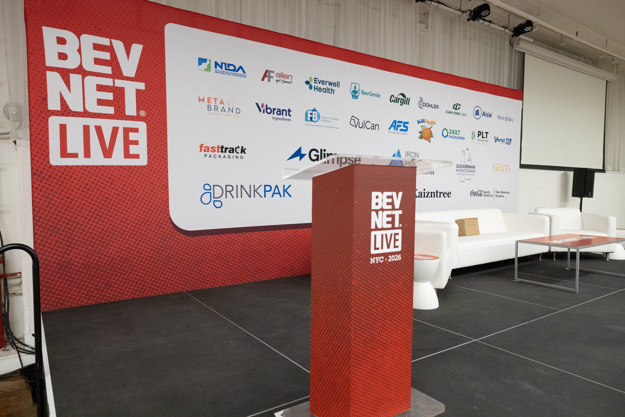 Stage with a red BevNET Live podium, white couches and a wall of sponsor logos beside a large BevNET Live panel.