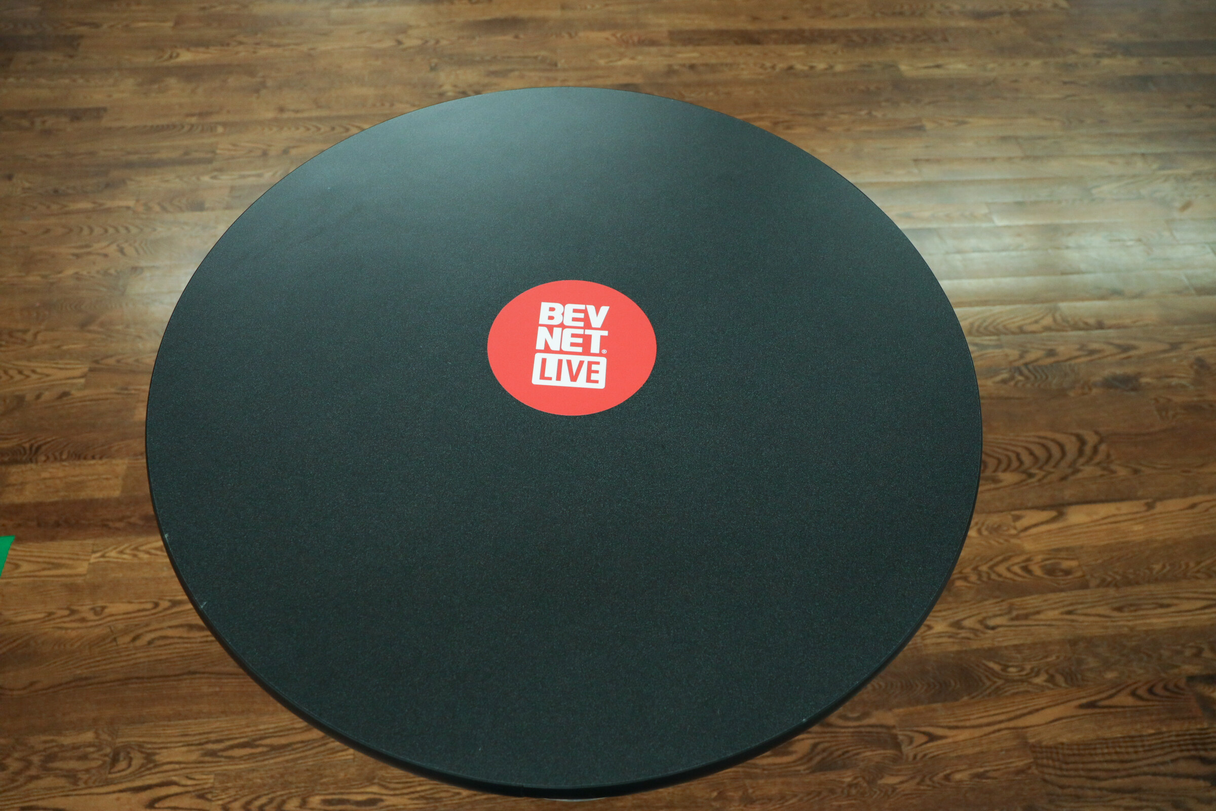 Overhead view of a round black table top with a red BevNET Live logo on a hardwood floor.