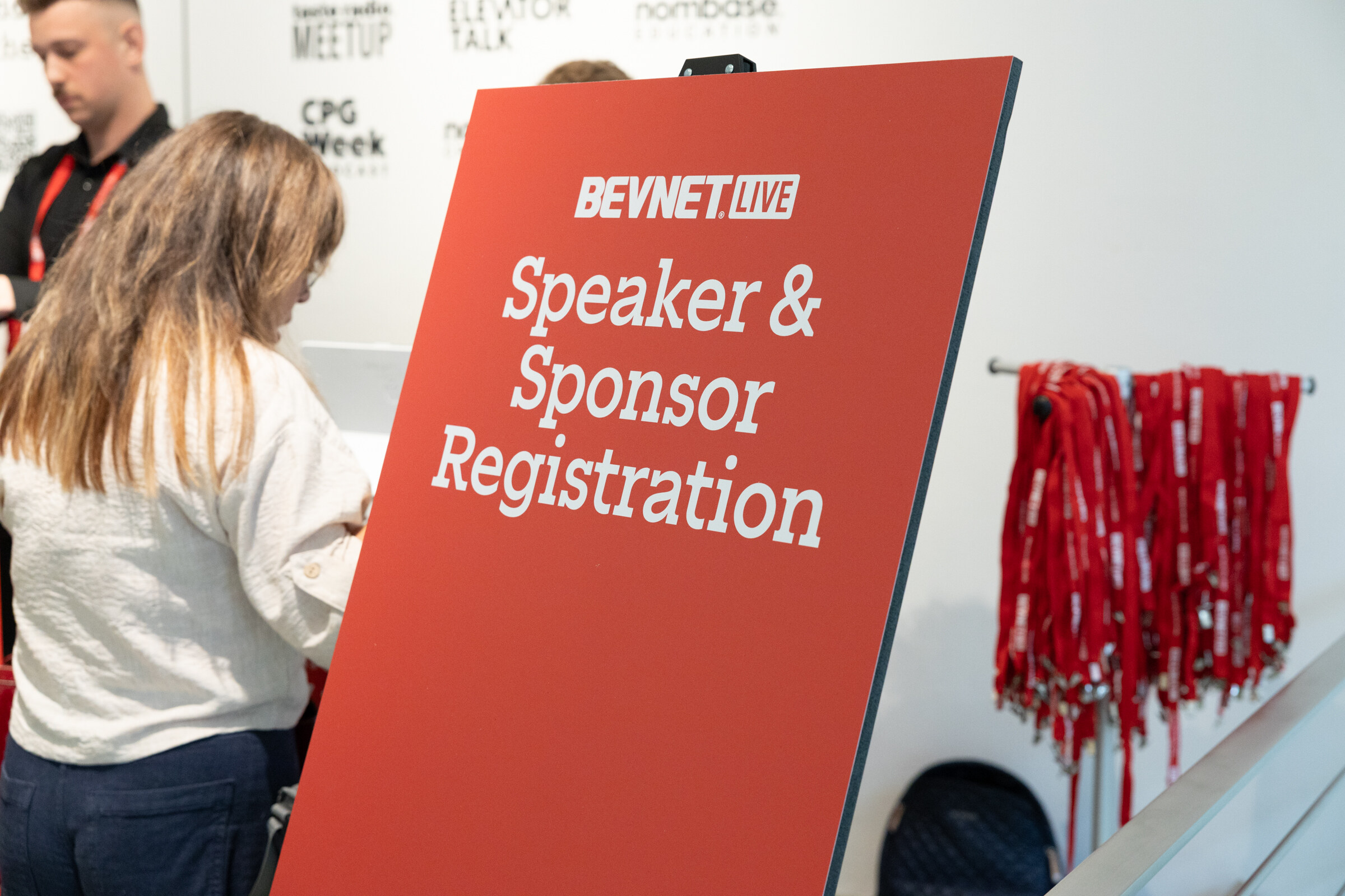 Red sign reading BevNET Live Speaker & Sponsor Registration beside a staff member and a rack of red lanyards.