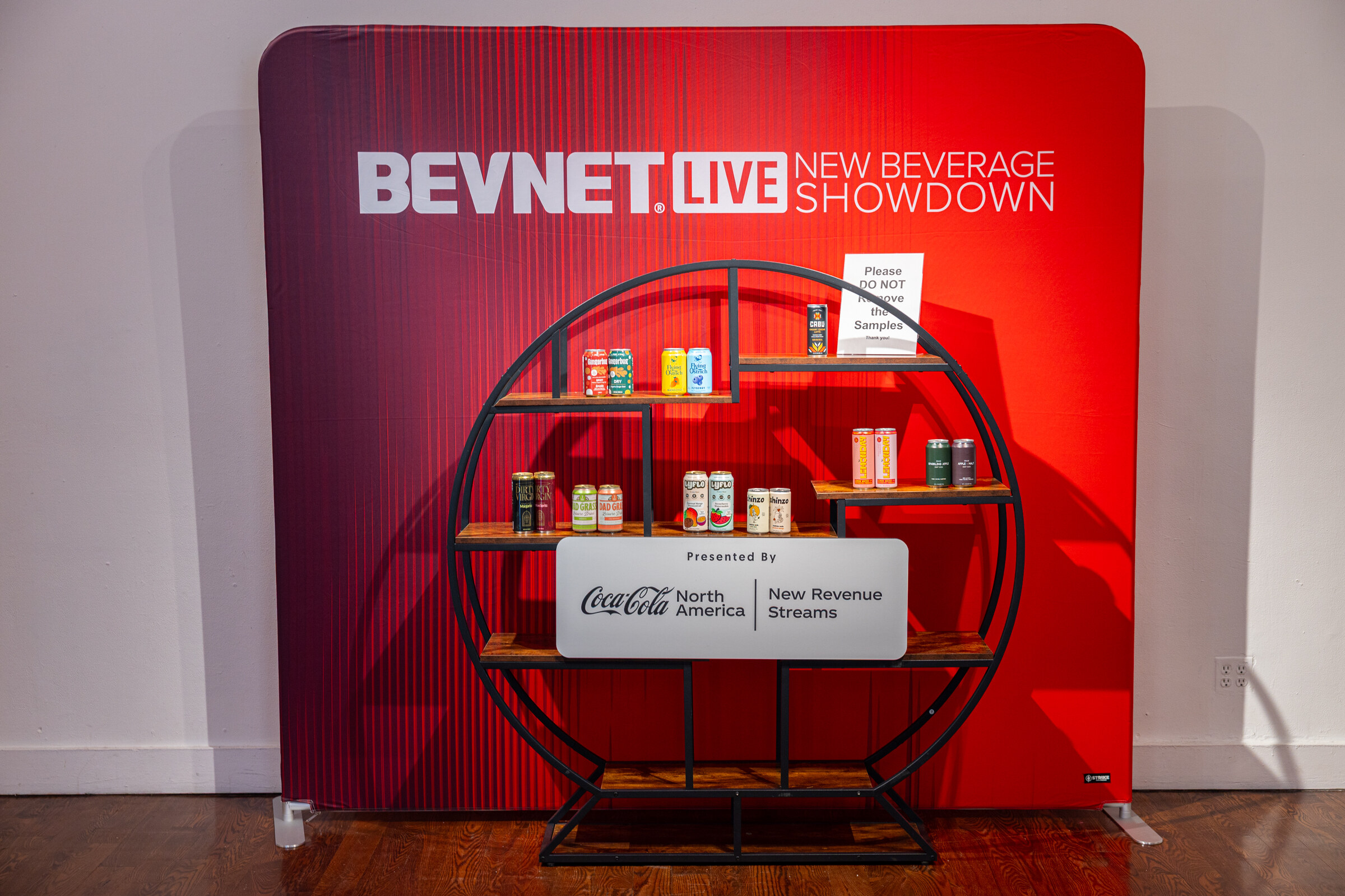 Red New Beverage Showdown backdrop with a round shelving unit of products, presented by Coca-Cola North America New Revenue Streams.