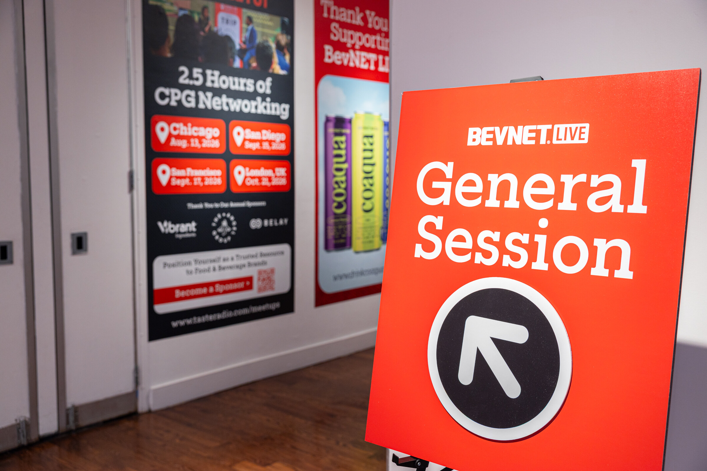 Red sign reading BevNET Live General Session with an arrow, in front of a wall of event posters.
